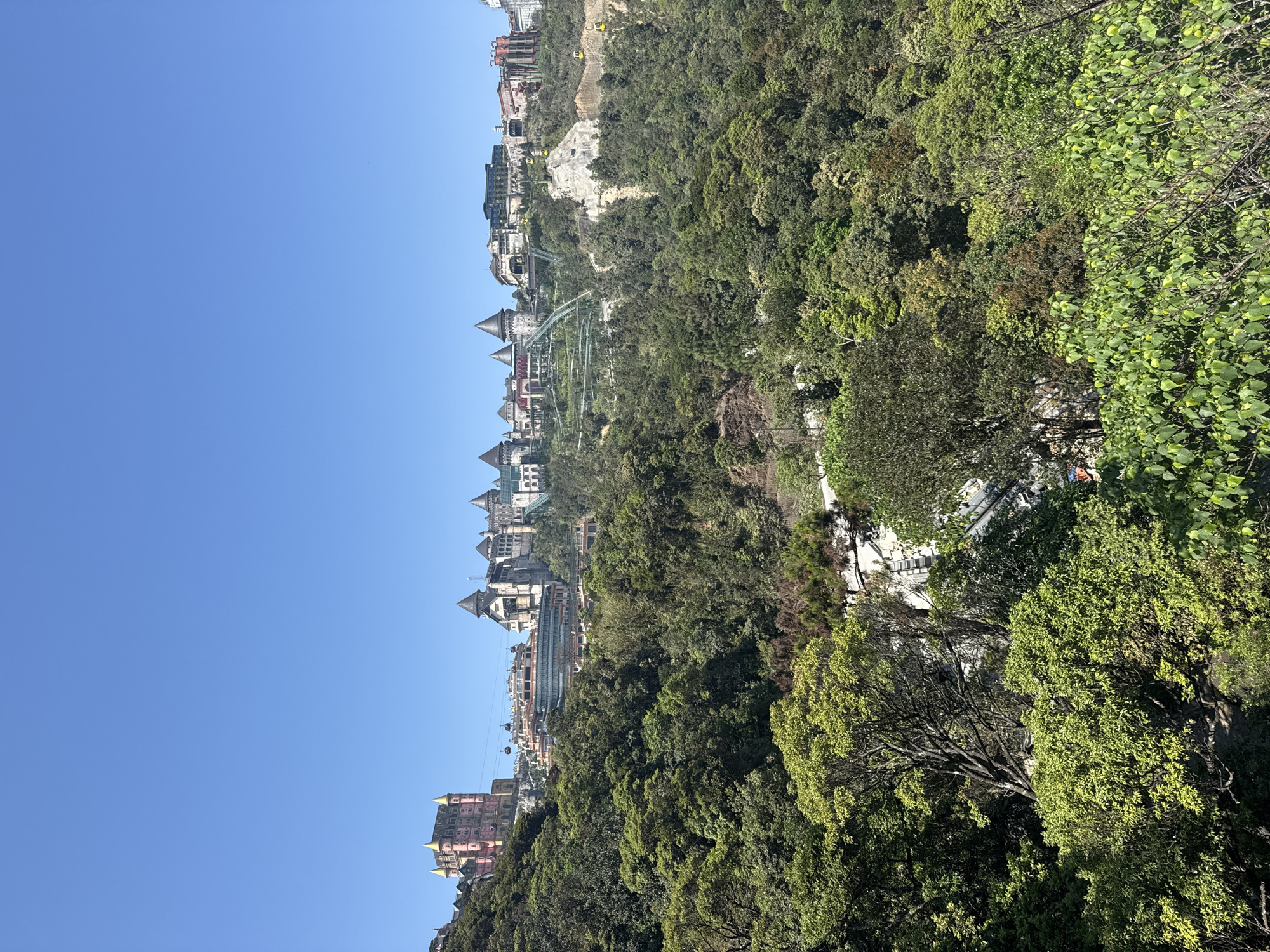 The image shows a view of Ba Na Hills in Đà Nẵng, Vietnam. Several buildings with European-style architecture are visible on a hill, surrounded by lush green trees. A roller coaster track and cable car lines are also seen winding through the landscape. The sky is clear and blue.