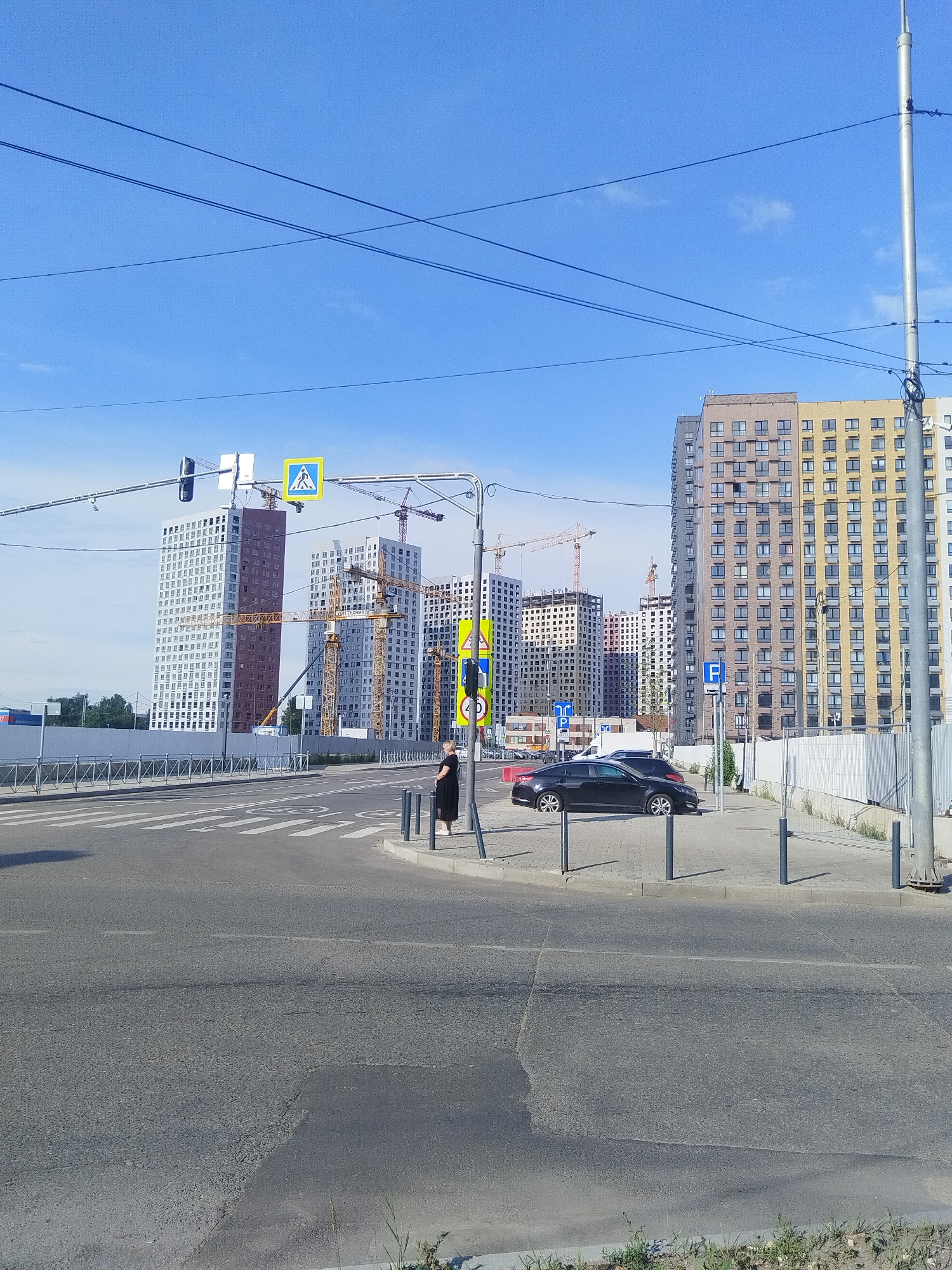 The image captures a street scene in Mytishchi, Russia, characterized by a bright blue sky and a mix of modern architecture and urban infrastructure. In the foreground, a paved road features pedestrian crossing markings and a slightly cracked surface in the lower half. Two black cars are parked on the right side of the road, near a sidewalk with bollards.
A woman in a black dress stands on the sidewalk next to a pedestrian crossing. Across the street, a series of tall, multi-story buildings are under construction, indicated by the presence of several yellow construction cranes. Some of these buildings are nearly complete, showcasing their facades with numerous windows, while others are still in earlier stages of construction. A few finished buildings with yellow and beige facades are also visible.
Traffic lights and road signs are present, including a pedestrian crossing sign and a speed limit sign. Overhead, power lines crisscross the sky. The overall environment suggests a developing urban area with ongoing construction projects.
