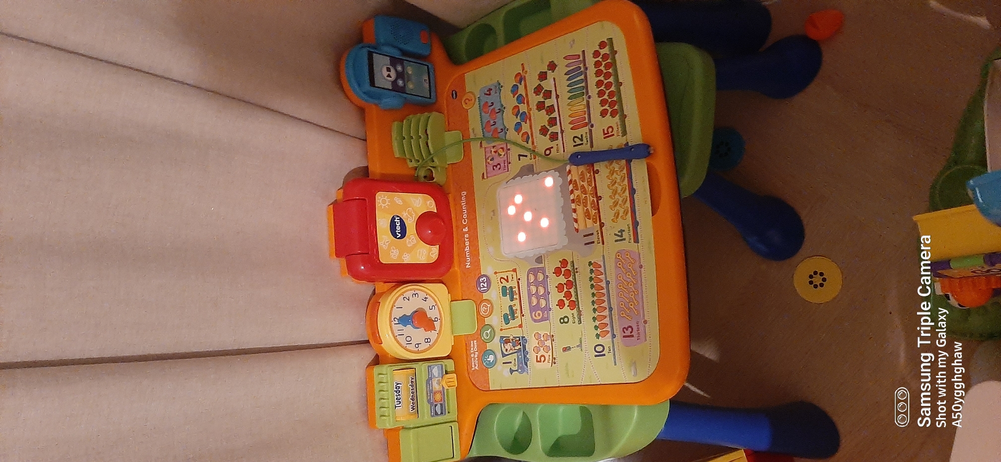 A VTech Numbers & Counting learning toy is displayed on a light brown carpeted surface, possibly a floor, in front of a tan-colored fabric backdrop. The toy is orange and green and features various educational elements, including a clock with movable hands, a digital display with illuminated dots, and illustrations of objects corresponding to numbers one through fifteen. A blue cord hangs from the toy. In the lower portion of the image, the bottom of the toy's legs are visible, along with parts of other toys and a yellow circular object on the carpet. The Samsung Triple Camera watermark is present in the bottom left corner. The image is a still photograph taken indoors, and no specific location in Dubai, United Arab Emirates, can be determined from the visual information provided.