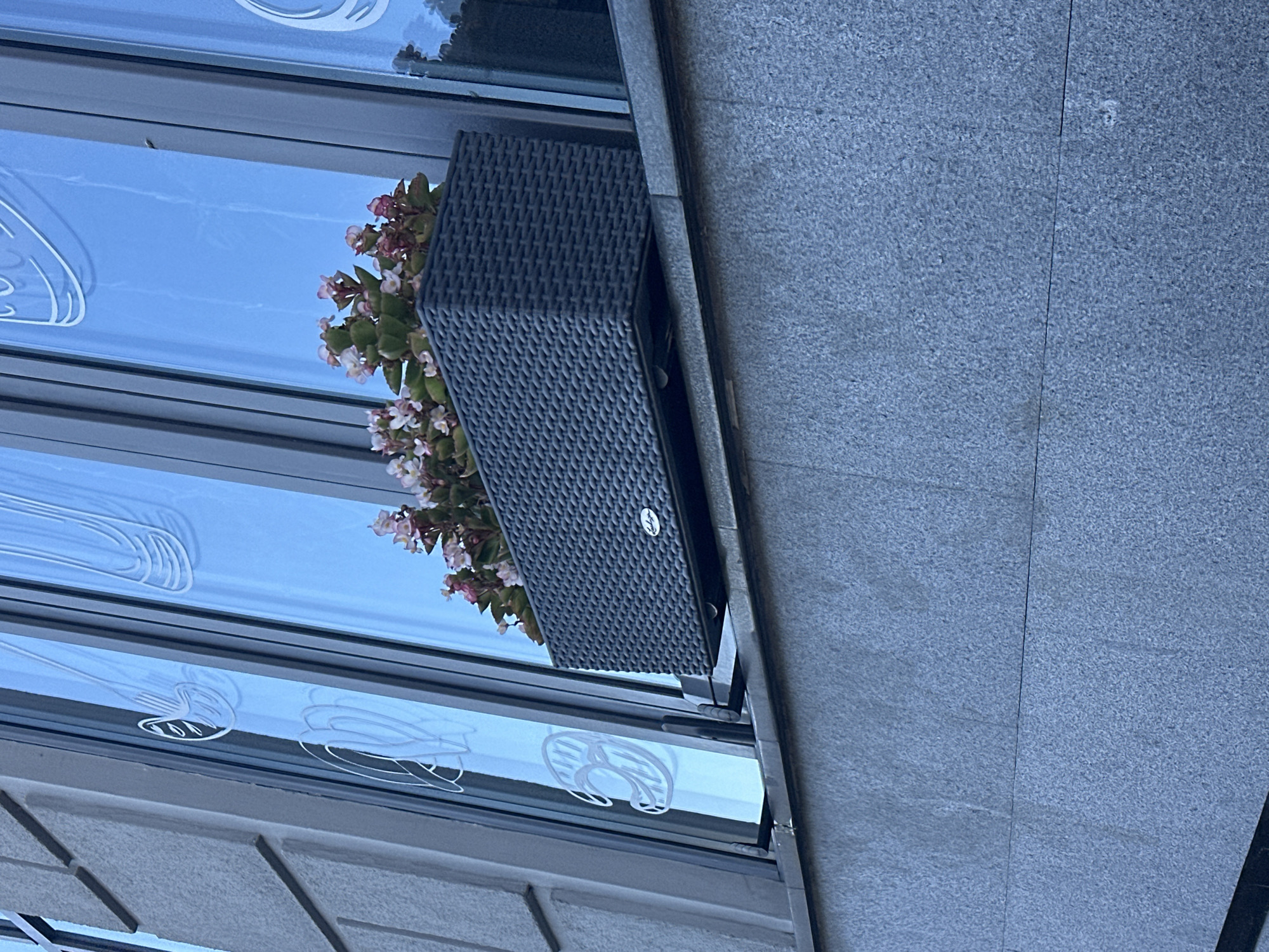 This is a low-angle, close-up, outdoor, eye-level shot of a planter box filled with small pink and white flowers. The planter box is dark grey, rectangular, and appears to be made of a woven material. It sits on a ledge outside a building, in front of tall, narrow windows. The windows have white decorative decals on them that appear to be abstract designs. The building facade to the left of the windows is made of textured grey stone blocks. The lower part of the image shows a similar textured grey stone surface, likely the building's exterior wall or a lower ledge. The overall lighting suggests it is daytime, possibly late afternoon, with soft natural light reflecting off the windows. There are no people visible in the image. The scene appears to be in an urban environment.