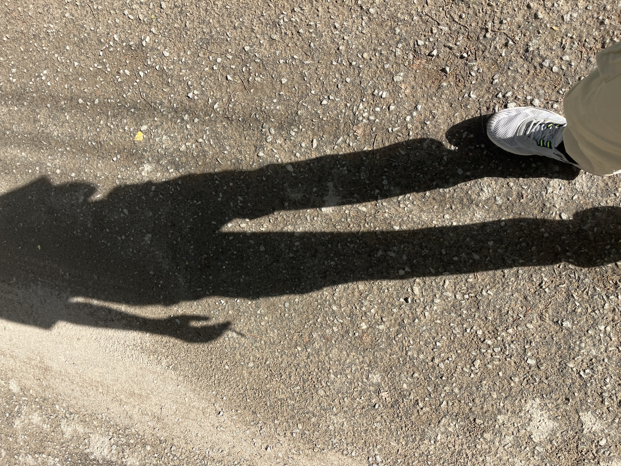 The image shows a person standing on a gravel and concrete surface. The shadow of the person is visible, with one arm outstretched. Part of the person's right foot, wearing a white sneaker with grey and green accents, is also visible. The location cannot be determined from the image.