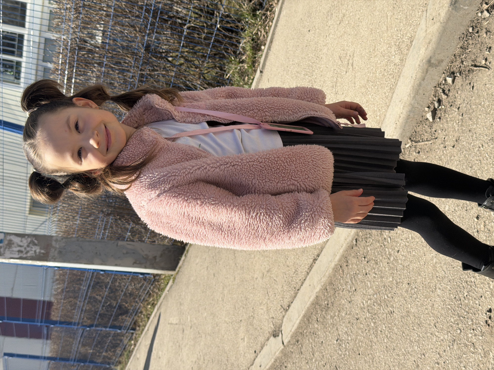 A young girl stands on a sidewalk alongside a road, next to a metal fence, in Perm, Russia. She wears a pink, fuzzy jacket, a white shirt, a pleated black skirt, black tights, and black shoes. Her hair is styled in two buns, and she is smiling. The sidewalk is concrete, and there is a curb between the sidewalk and the road. The road surface appears to be made of a light-colored material. A building is visible behind the fence. It is a sunny day.