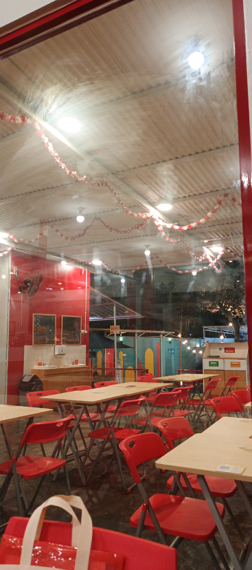 The image depicts an indoor dining area, likely a restaurant or cafe, with an array of tables and red chairs. The scene is observed from outside, through a glass pane, as indicated by reflections on the surface. The interior features a corrugated ceiling adorned with red and white festive streamers and illuminated by overhead lights.
On the left side of the image, a bright red wall is visible, with two mirrors mounted above a sink and countertop area. A black trash bin is situated next to the sink. Further into the interior, colorful panels in green and yellow form a backdrop, with string lights illuminating the space.
The foreground is dominated by several light-colored tables with metal folding legs, surrounded by numerous red plastic chairs. In the lower left corner, a white and red bag is placed on a red surface, possibly a chair or table.
The image was taken at a restaurant located in Sidoarum, Indonesia. The time of day appears to be evening or night, given the ambient lighting and the darkness visible outside through the interior spaces. No people are clearly visible within the dining area.