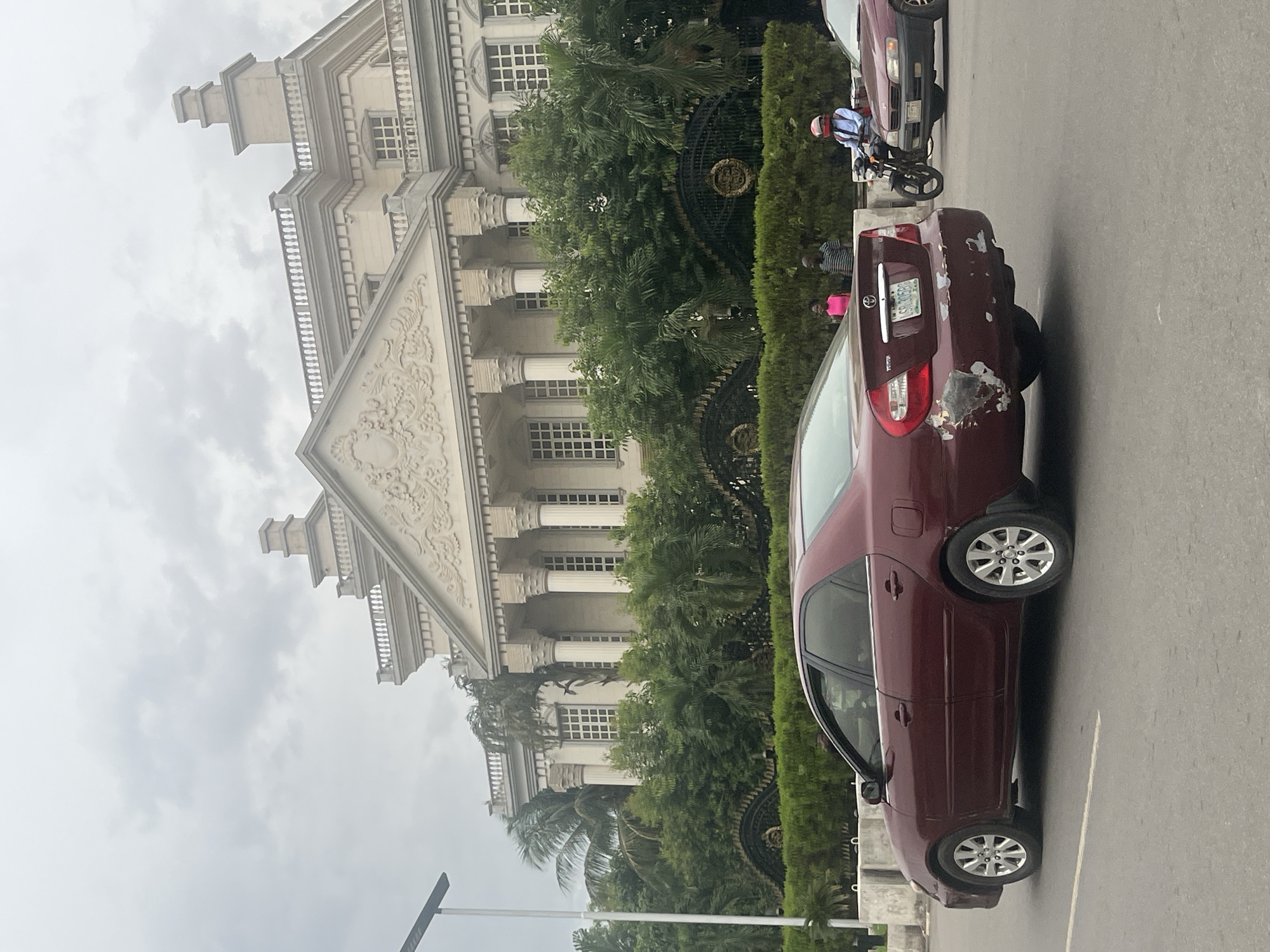 The image shows a residential street in Abuja, Nigeria, in front of a large, ornately decorated building with many windows and columns. The building is set behind a tall green hedge and a decorative wrought-iron fence. On the right side of the street, a maroon Toyota car is parked. The car appears to have damage on the rear fender and bumper. Next to the maroon car, a second car is partially visible. A person on a motorcycle is stopped on the street between the two cars. A few people stand near the maroon car. The sky is overcast.
