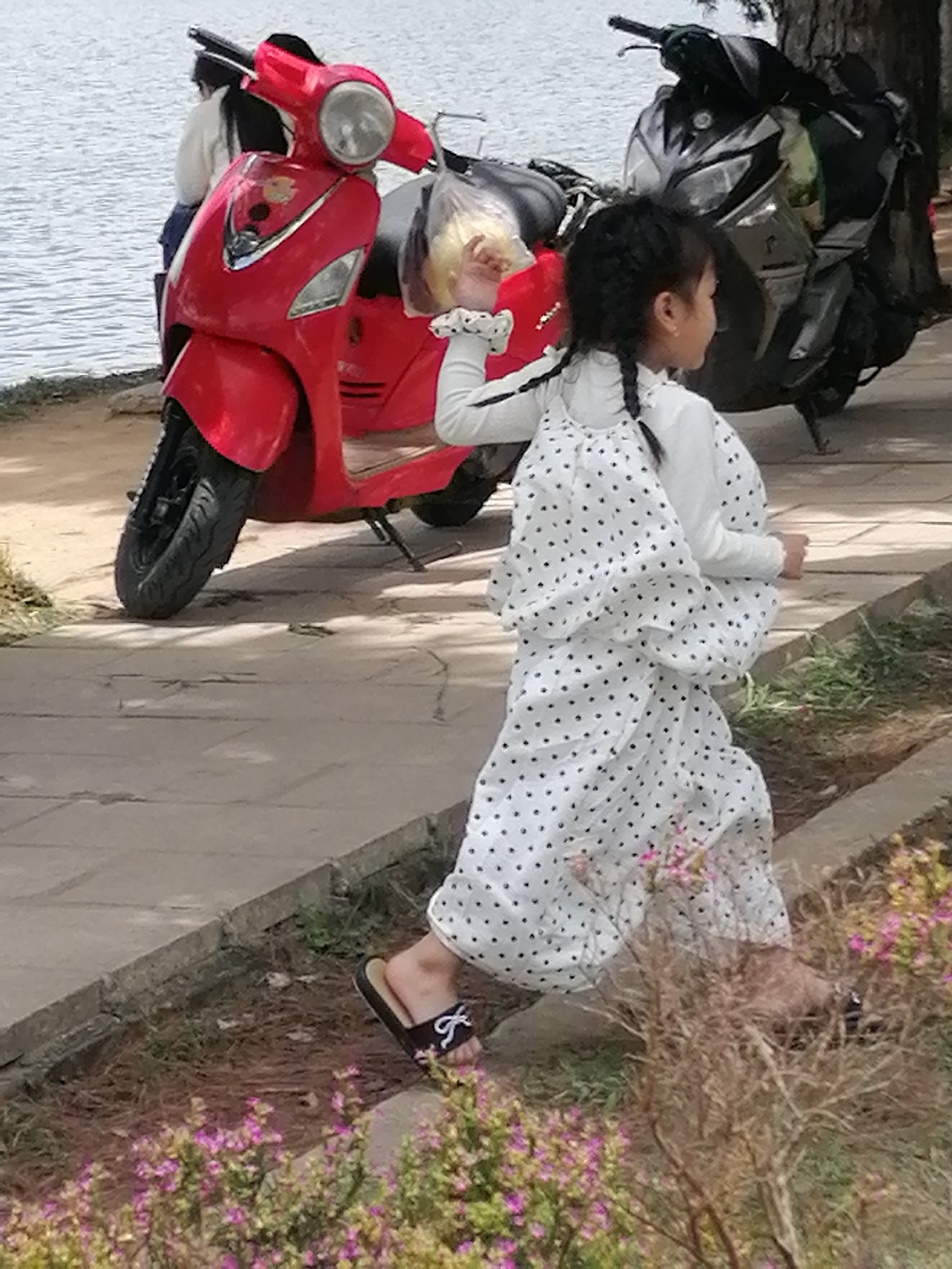 In Da Lat, Vietnam, a young Vietnamese girl with braided hair, wearing a white polka-dot dress and dark sandals with bows, is captured mid-stride on a paved walkway beside a body of water. Her body is angled away from the viewer, with her head turned to the right as she runs. She holds a plastic bag containing yellow objects in her right hand. Behind her, a red scooter is parked on the walkway. Further in the background, a black scooter is visible, and beyond that, the glistening surface of a lake stretches out under an overcast sky. The scene is bordered by a mix of dry and green vegetation.