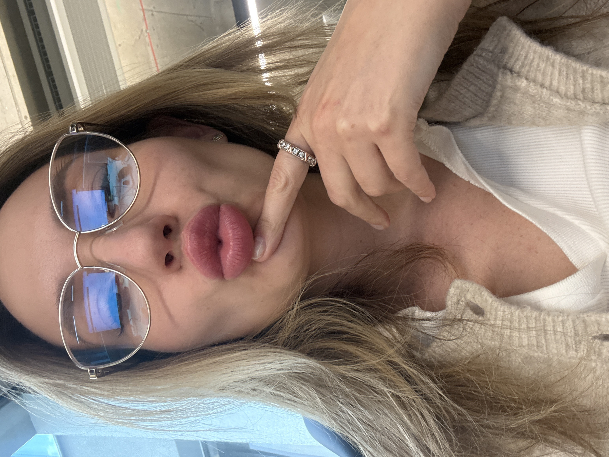 A woman with blonde hair is wearing glasses and a ring. She is making a duck face while holding a finger under her lip. The background is blurry and suggests an indoor setting. There is no way to verify that this is in Riga, Latvia.