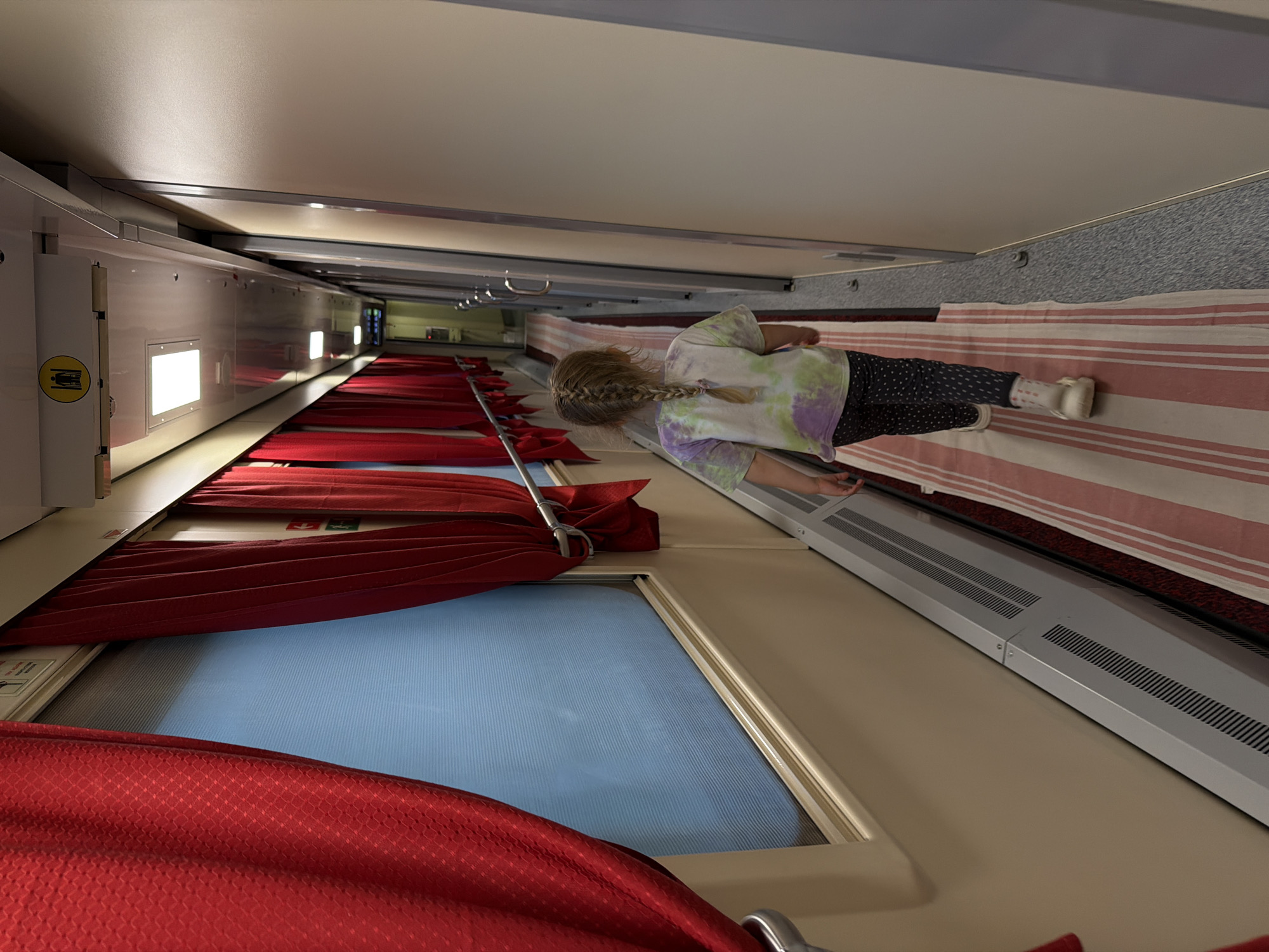 The image shows the interior of a train car, specifically a couchette car, in an unknown location. The viewpoint is looking down the length of the car, showing rows of berths on one side. A young girl is standing in the aisle, facing away from the camera. She is wearing a tie-dye shirt and patterned leggings. The berths have red curtains partially drawn, revealing windows with a view of the outside. The upper berths appear to have striped bedding. The interior is clean and well-lit.