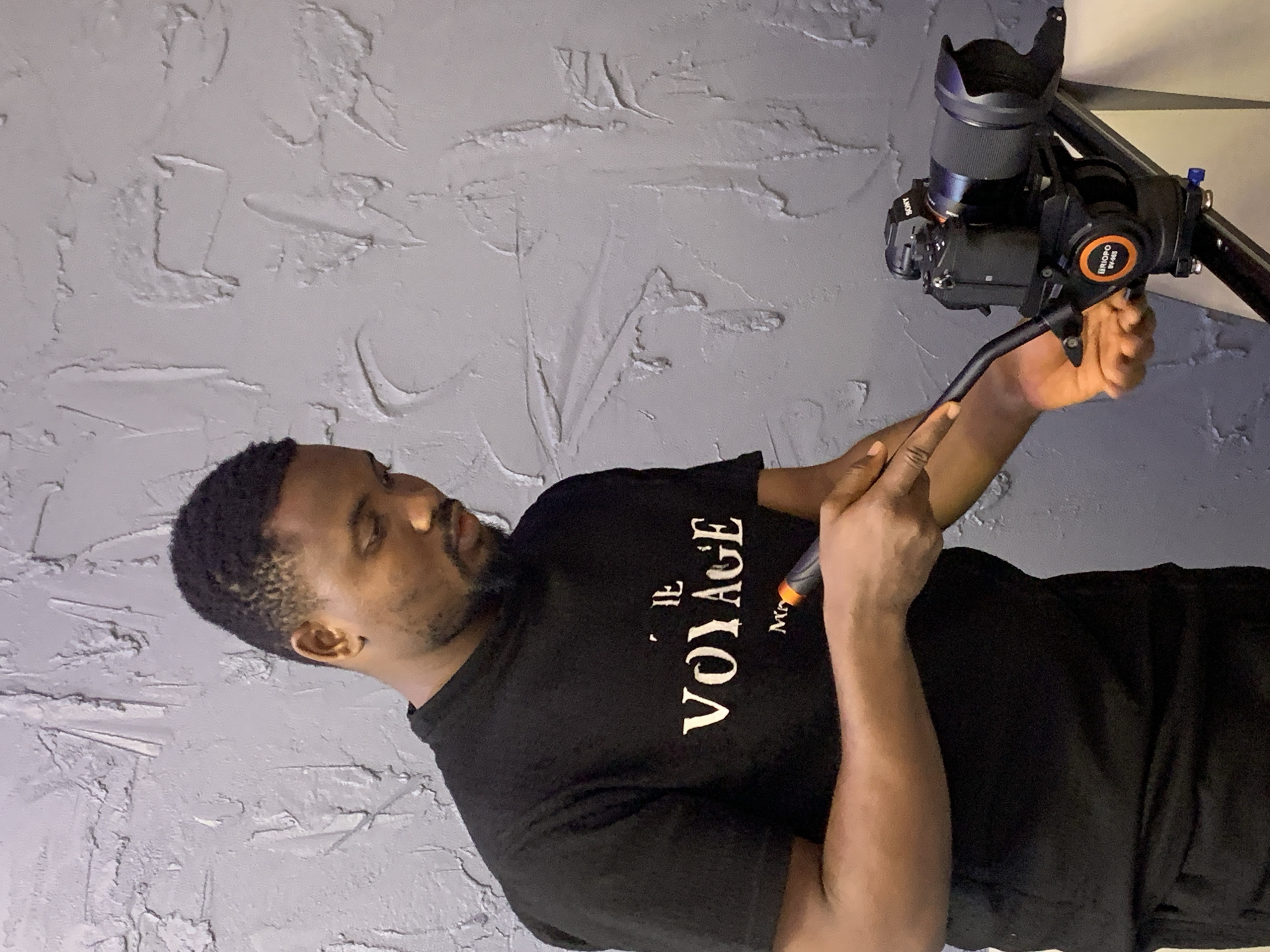 An adult male is observed indoors in Igboefon, Nigeria, positioned against a grey, textured stucco wall. He wears a black short-sleeved t-shirt bearing the white inscription "LE VOYAGE". The individual has short dark hair and a beard, with his gaze directed downwards towards the equipment. He is engaged with a professional video camera setup, holding a black handle with an orange tip in his right hand. His left hand is positioned near the base of a black Sony camera, which is mounted on an orange-branded "RIIOPO BV-66" fluid head. The fluid head is affixed to a dark, track-based camera support system, partially visible in the lower right of the frame. The scene depicts the operation or adjustment of video production equipment.
