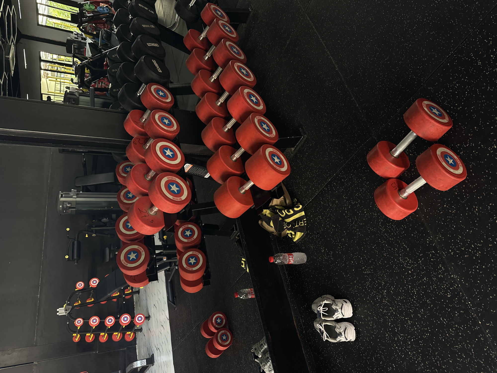 The image captures a gym scene in North Kuta, Indonesia. Red dumbbells with a Captain America shield design are displayed on racks and scattered on the black speckled floor. There is also a set of black dumbbells on a rack nearby. A yellow and black towel, two water bottles, and two pairs of sneakers are on the floor near the dumbbells. The gym setting is suggested by the presence of exercise equipment and weights, under window lighting.