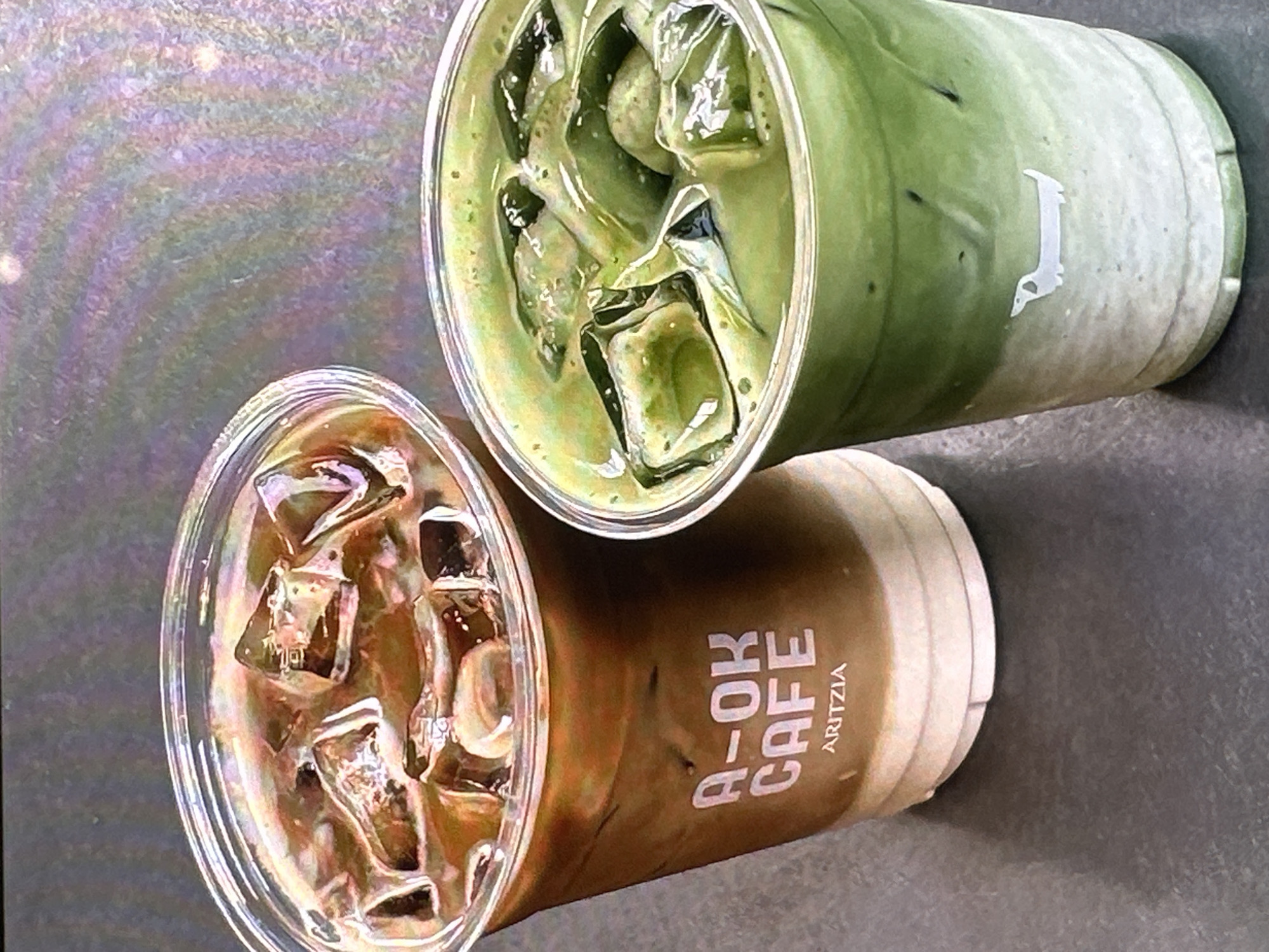The image shows two cups of iced beverages. The cup on the left contains a brown liquid with ice cubes, with the text "A-OK CAFE ARITZIA" visible on its side. The cup on the right contains a green liquid with ice cubes and displays a white logo of a goose standing on one leg. The drinks are positioned on a grey surface with a slight sheen. The background is blurred, suggesting a shallow depth of field. The setting appears to be a cafe or casual dining area, potentially in Yerevan, Armenia, due to the Aritzia brand, which has locations in Armenia, and the presence of the A-OK CAFE. The lighting seems natural.