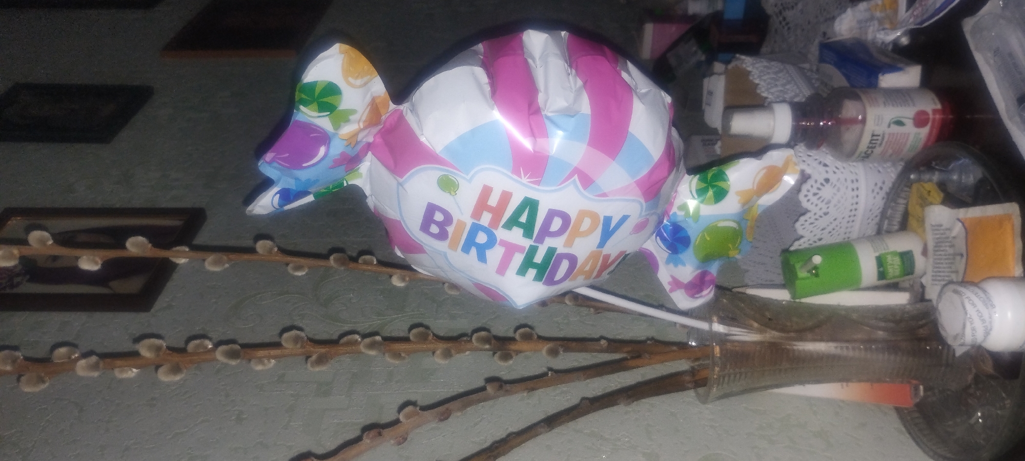 The image displays a still life composition that appears to be indoors. The primary subject is a large, candy-shaped balloon with the words "HAPPY BIRTHDAY" printed on it in colorful letters. The balloon has a pink and white striped pattern and is decorated with images of various candies. In front of the balloon, several branches of pussy willows are arranged in what looks like a vase, though the vase itself is mostly obscured. The background is cluttered with various objects, including bottles, boxes, and decorative lace doilies, suggesting a shelf or tabletop. Some of the bottles appear to be medicinal or cosmetic. There are also framed pictures visible on the wall in the background. The lighting is dim, casting shadows and giving the scene a somewhat muted appearance. The overall impression is one of a casual indoor setting, possibly a home, with a festive element introduced by the birthday balloon. The pussy willows suggest the possibility of spring. No people are visible in the image.