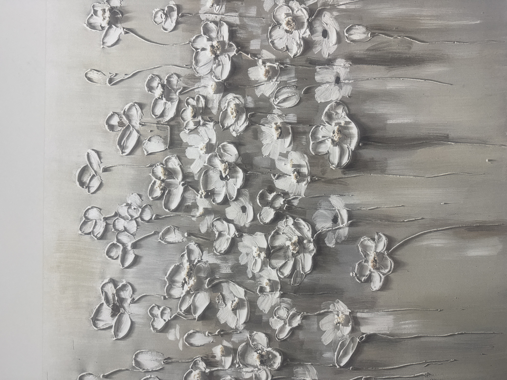 The image displays a detailed view of a three-dimensional floral artwork, primarily rendered in a monochromatic scheme of white and various shades of grey. Numerous white flowers and flower buds are depicted in high relief, sculpted with thick impasto strokes that give them a palpable, tactile quality. Many of the open flowers feature subtle light golden-brown or beige accents in their centers, suggesting stamens.
The background of the artwork consists of textured horizontal brushstrokes in light to dark grey tones, providing a muted yet dynamic backdrop. Thin, raised lines, resembling delicate stems or vines, connect the individual floral elements, weaving across the textured grey surface. The flowers are arranged in an organic flow, appearing to emerge from the bottom left and spreading diagonally and vertically across the canvas, creating a sense of natural movement despite their static nature.
The strong relief of the white flowers casts noticeable shadows, further enhancing their three-dimensionality and creating depth within the piece. The overall aesthetic is elegant and modern, emphasizing texture, form, and a serene color palette. No human subjects are present, and no specific activity or event is taking place beyond the visual representation of the artwork itself. There is no visible text.