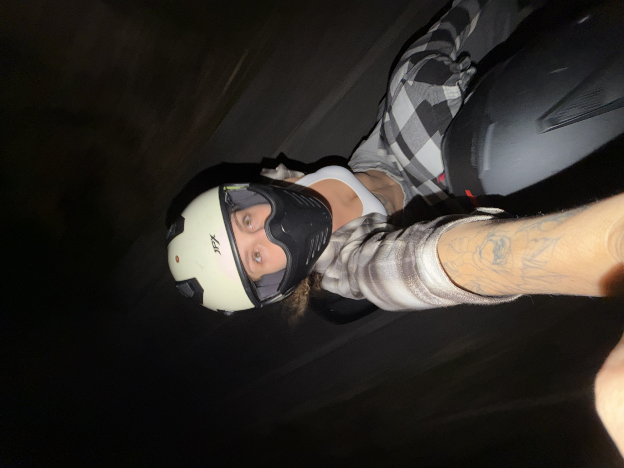 The image displays a close-up, angled selfie of a person at night, likely taken outdoors in North Kuta, Indonesia. The individual is wearing a cream-colored motorcycle helmet with a black visor or face mask; the brand "JPX" is visible on the side of the helmet. Their eyes, with light irises, are visible through the helmet's opening, looking directly at the camera with a composed expression.
The person is dressed in a black and white/grey plaid long-sleeve shirt worn open over a white tank top. Tattoos are discernible on their visible arm. To the right of the frame, a dark, indistinct object, possibly part of a motorcycle or vehicle, can be seen. The background is largely dark and blurred, suggesting motion or very low light conditions, indicative of a nighttime setting. The composition and attire imply the person is either riding or posing on a motorcycle during an evening activity.