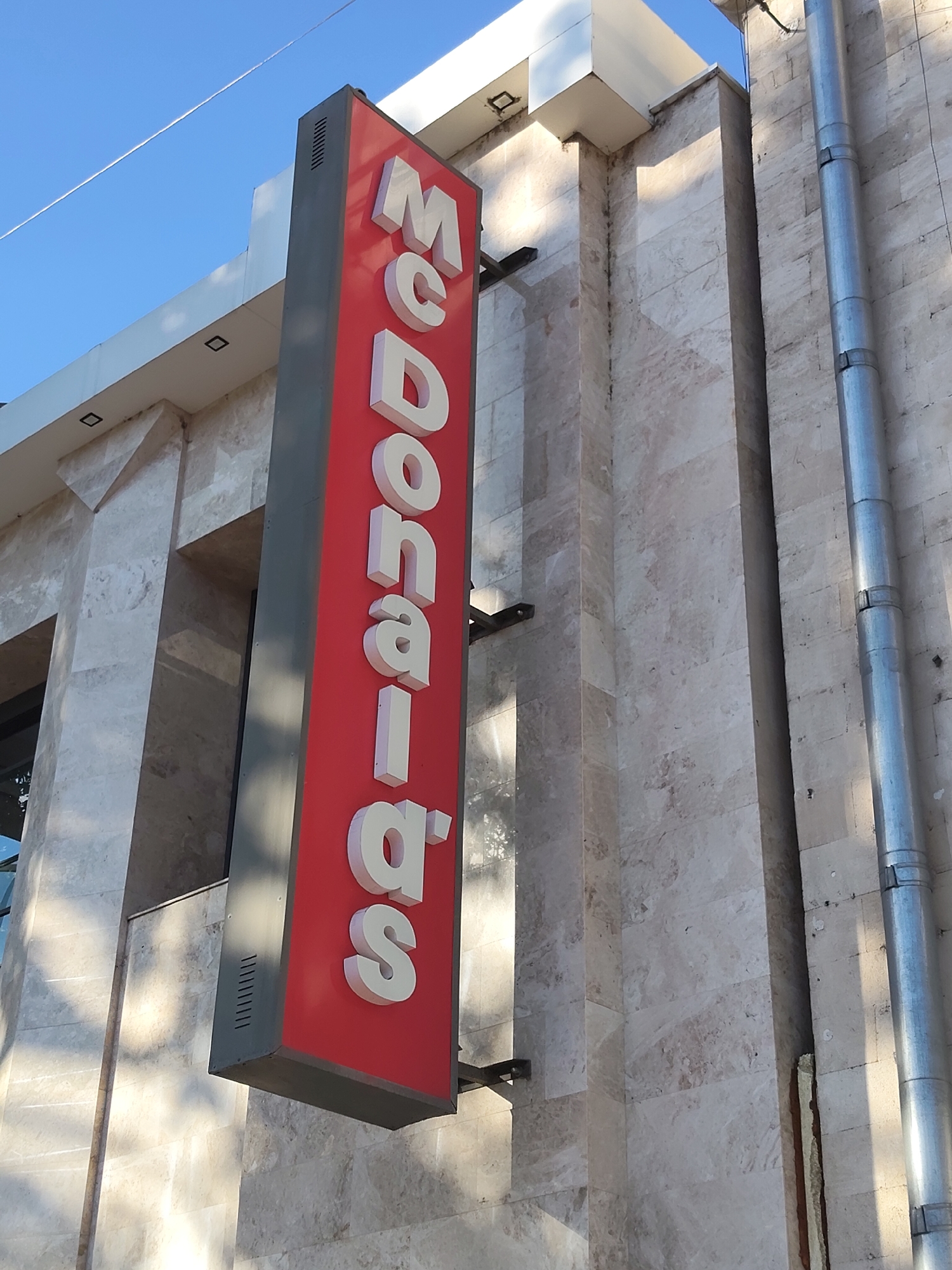 An outdoor vertical sign for McDonald's hangs on the side of a light-colored stone building. The sign is red with large white letters spelling out "McDonald's" vertically. The building has a modern design with large rectangular sections of stone and some white architectural elements at the top. A silver downspout runs vertically along the right side of the building. The sky above is clear and blue with a thin white line, possibly a contrail, streaking across it. The lighting suggests it is daytime, with shadows cast on the building. The image was taken in Chișinău, Moldova.