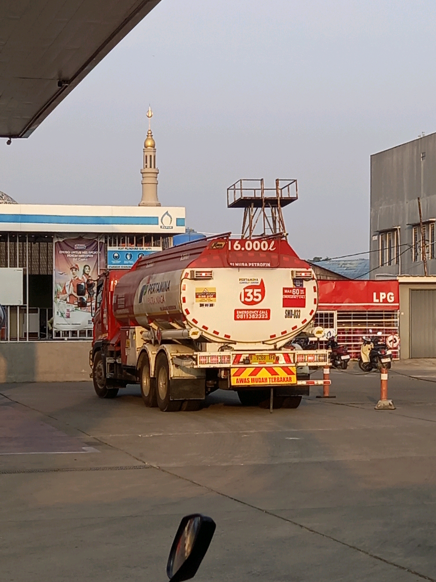 This image captures a tanker truck with "PERTAMINA" branding, suggesting it's associated with the Indonesian state-owned oil and gas company. The truck is parked at what appears to be a gas station or a refueling facility. In the background, a mosque with a golden dome is visible, along with a building labeled "LPG". The overall scene depicts a typical urban or semi-urban environment in Indonesia, likely Samarinda given the context. The tanker truck is the primary subject, occupying the central portion of the frame. The surrounding environment includes paved ground, a canopy structure, and other vehicles, indicating a place of commerce or industry.
