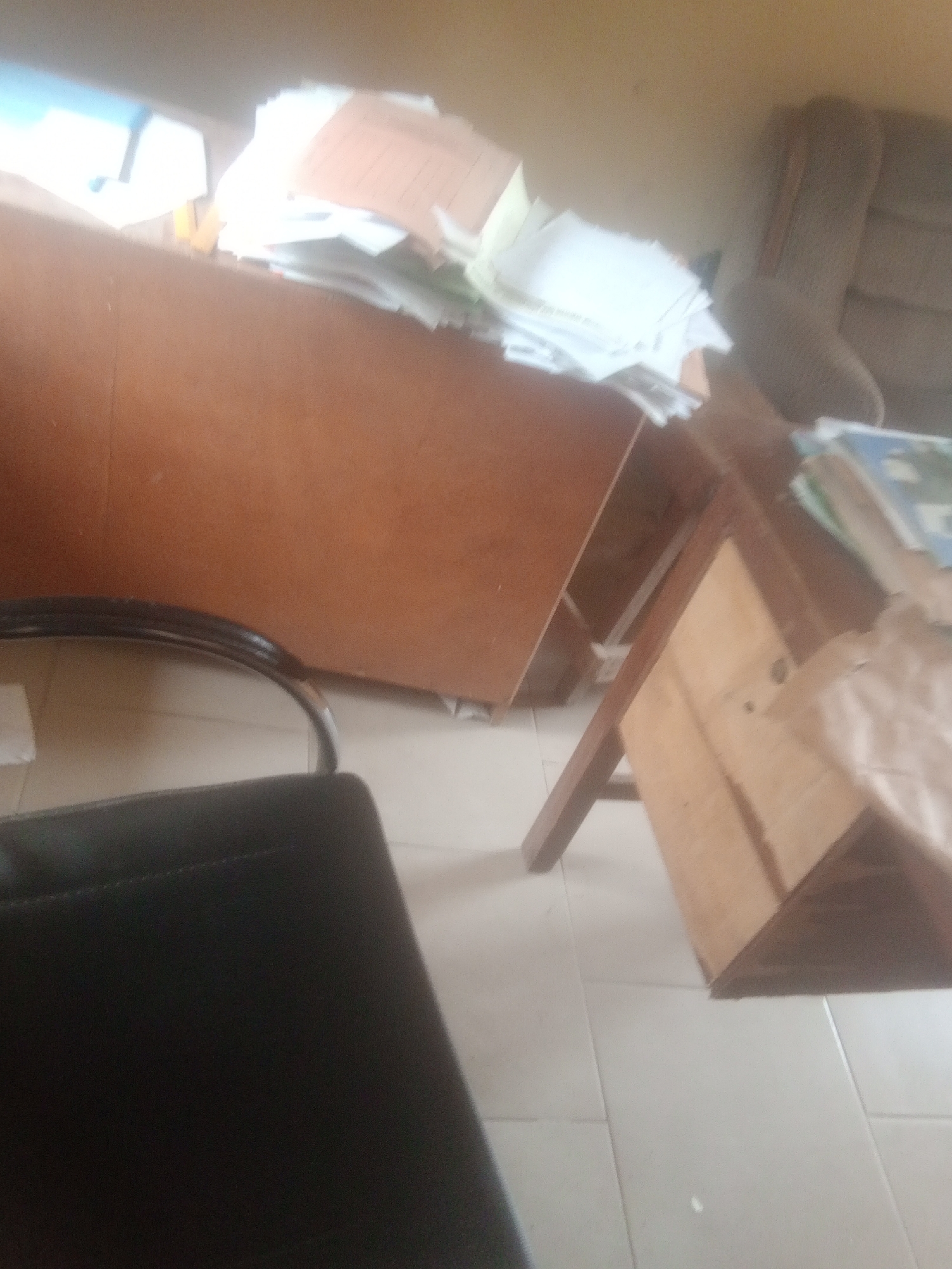 This is a medium-shot, interior photograph taken in Jalingo, Nigeria. The scene depicts an office environment, possibly a home office given the informal arrangement. The primary focus is on a wooden desk and a large wooden cabinet filled with papers. The cabinet is overflowing with documents, suggesting a busy or perhaps disorganized workspace.
To the left of the frame, a black office chair is partially visible, with its armrest in focus. In the background, a beige armchair is situated, its texture suggesting fabric upholstery. The lighting in the room appears to be from overhead sources, creating a bright, slightly washed-out effect, particularly on the piles of paper.
The desk itself is made of wood and has a drawer. It is positioned to the right of the cabinet. On the desk, more papers and some books are stacked, with one book showing a blue cover. The floor is tiled in a light color, typical of many interior spaces. The overall impression is one of an occupied and active workspace, though no people are directly visible. The image quality is somewhat blurry, potentially due to movement during capture or low resolution, which obscures finer details. There is no discernible text visible in the image. The time of day is difficult to ascertain definitively, but the bright interior lighting suggests it is daytime.