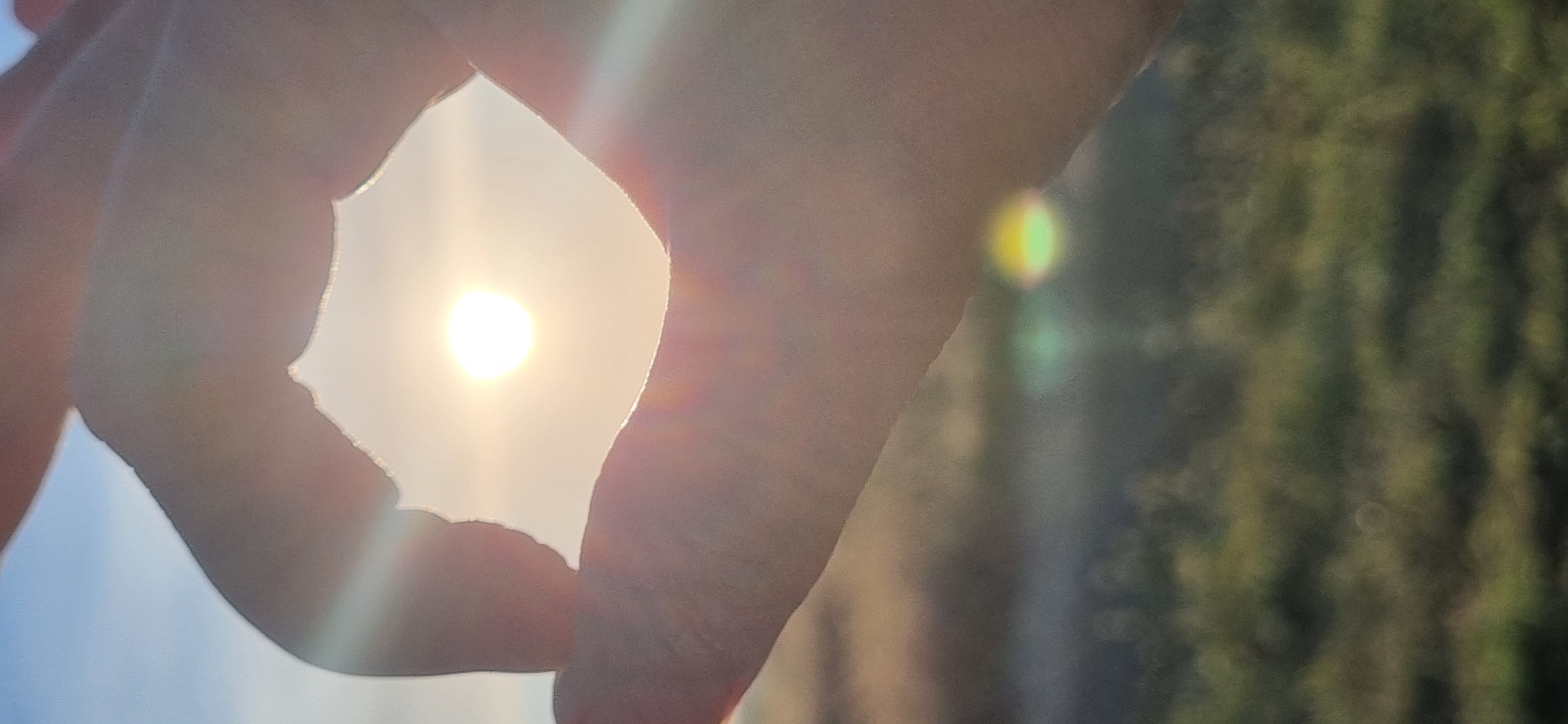 The image shows a person's hands framing the sun. The sun is bright and appears to be setting or rising. The background is blurry and appears to be a natural landscape with some greenery. There are lens flares visible, indicating a strong light source. The image was taken in Blizhnee, Ukraine.