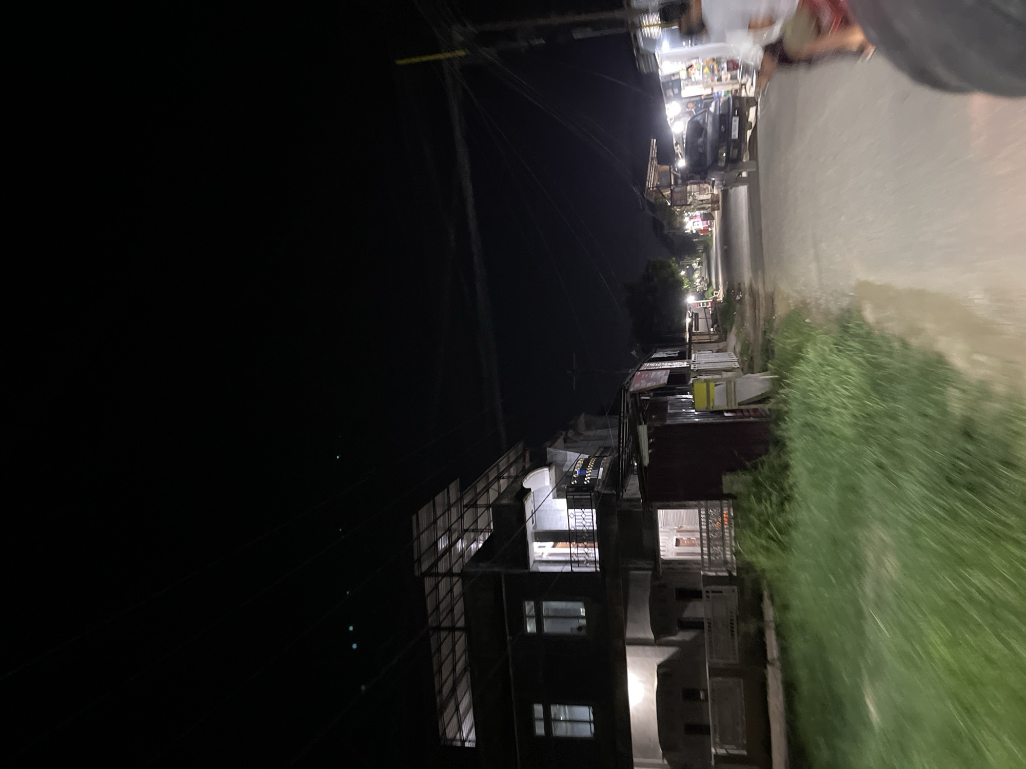 The image depicts a nighttime street scene in the city of Tanjungkarang, Indonesia. A multi-story building with illuminated windows and a metal roof structure stands on the left side of the frame, bordered by a patch of tall grass. The street itself is visible on the right, with a dark-colored SUV driving away from the camera. Along the street, there are several small shops or stalls with lights illuminating their fronts. A person is partially visible in the foreground on the right, suggesting the photo was taken from a moving vehicle, possibly a motorcycle. The sky is dark, indicating it is nighttime, and power lines are visible overhead.