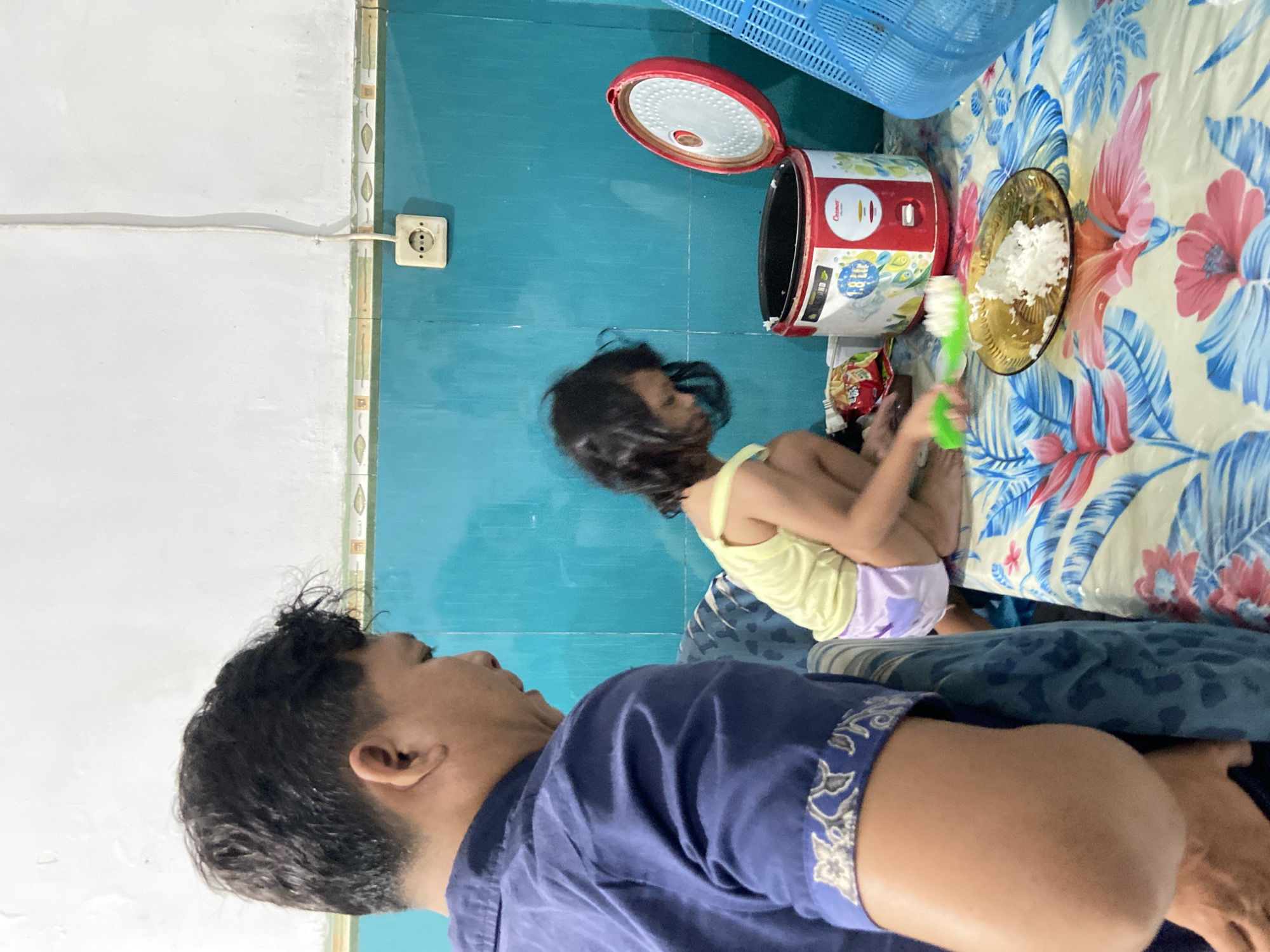 The image depicts a domestic scene in a room, likely a kitchen or dining area, located in the city of Gelam, Indonesia. In the foreground, a man wearing a blue shirt is partially visible from the side, facing away from the camera. He appears to be an adult male with dark hair. To the right of the man and further into the scene, a young child with dark hair is crouching. The child is wearing a yellow tank top and purple shorts and is interacting with food items on a table.
The table is covered with a colorful, floral-patterned tablecloth. On the table, a rice cooker with its lid open is prominent. Beside the rice cooker is a plate with a portion of white rice, and the child is holding a green utensil, possibly a spoon or spatula, near the plate. A bag of snacks is also visible on the table. The background of the room features a turquoise-colored wall with a white electrical outlet and cord. A blue plastic basket is positioned on the right side of the image. The overall lighting suggests an indoor setting.