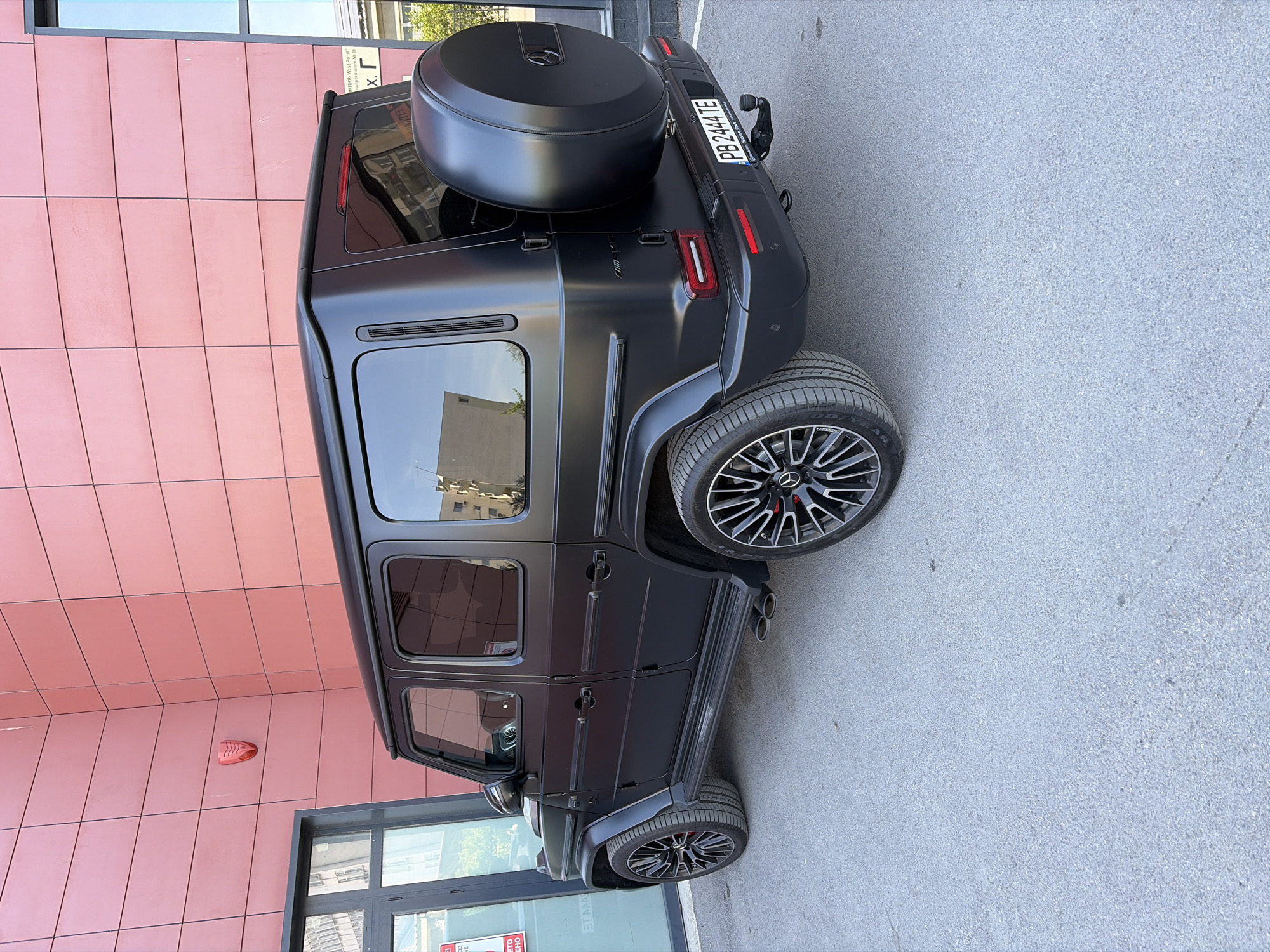 The image features a matte black Mercedes-Benz G-Class (G-Wagen) SUV, parked outdoors during the daytime under clear, sunny weather. The luxury vehicle is viewed from its rear-passenger side, displaying its robust design, black multi-spoke alloy wheels with visible red brake calipers, and side-mounted exhaust pipes. A black spare tire cover, embossed with the Mercedes-Benz logo, is prominently mounted on the rear door.
The SUV is stationary on a light grey asphalt surface, positioned alongside a contemporary building. This building boasts a distinctive salmon or terracotta-colored tiled facade, arranged in a grid pattern, and features large reflective windows. Reflections in the car's tinted windows show elements of surrounding architecture and greenery. A red alarm or siren unit is affixed to the building's facade.
The location is identified as Plovdiv, Bulgaria, confirmed by the license plate "PB 2444 TE" (where "PB" is the regional code for Plovdiv) and partially visible Cyrillic text on the building, including "Пловдив-West Point" and "Озерско шосе № 38." Other visible text includes "AMG" on the vehicle's rear and "GOOD YEAR" on the tire sidewalls. No people or active events are depicted.