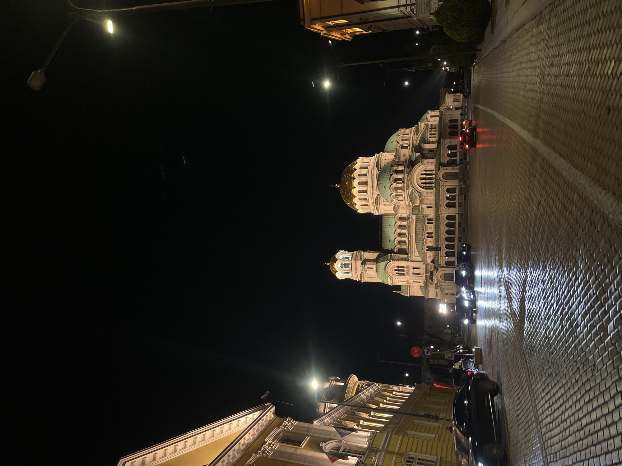 The image presents a nighttime street scene in Sofia, Bulgaria, dominated by the illuminated St. Alexander Nevsky Cathedral. The cathedral, a grand Orthodox edifice, stands prominently in the center-right of the frame, showcasing its white stone facade, multiple green-domed roofs, and distinctive golden central domes, all brightly lit against the dark sky.
A wide, cobbled street extends from the foreground into the distance, with a single white line running down its center. Several cars are visible on the street; some are parked along the left side, while one red car with illuminated taillights appears to be moving or stopped further down the road. Headlights from other vehicles cast reflections on the wet-looking pavement. On the right side of the street, more buildings with classical architectural elements and warm lighting line the thoroughfare.
Streetlights with bright, warm yellow glows illuminate the scene from above and along the street. The sky is completely dark, indicating deep night. On the right side of the road, a red "STOP" sign with white text is clearly visible, along with a blue circular sign next to it, likely indicating a traffic directive. The overall mood is serene and majestic, highlighting the architectural beauty of the cathedral at night.