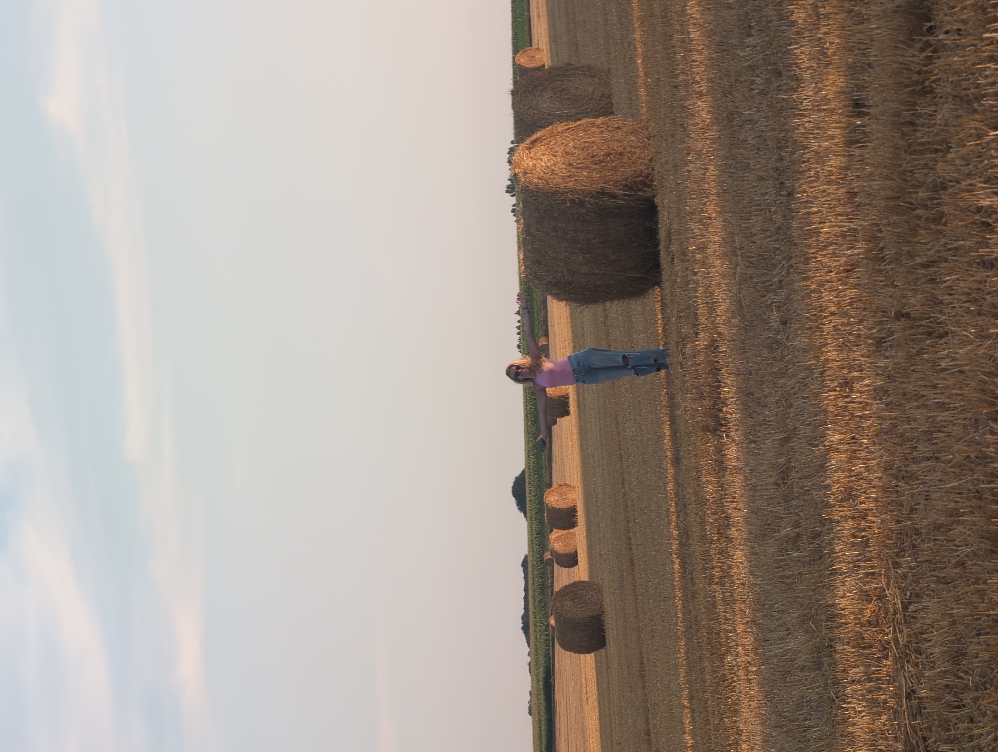 An outdoor, medium-shot photograph captures a woman standing in a harvested field with round hay bales scattered across the landscape. The woman, positioned slightly to the left of the frame's center, wears a pink tank top and ripped blue jeans. She is facing the camera with her arms outstretched to the sides and appears to be smiling, with sunglasses on her face. Behind her, several large, round hay bales are visible, most notably a prominent one to her right. The field is covered in dry, golden straw, with stubble from the harvested crop. In the background, a strip of green vegetation, possibly a field of corn or trees, separates the harvested field from the pale blue sky. The lighting suggests it is late afternoon or early evening, casting long shadows across the field. The location is identified as Alvimare, France.