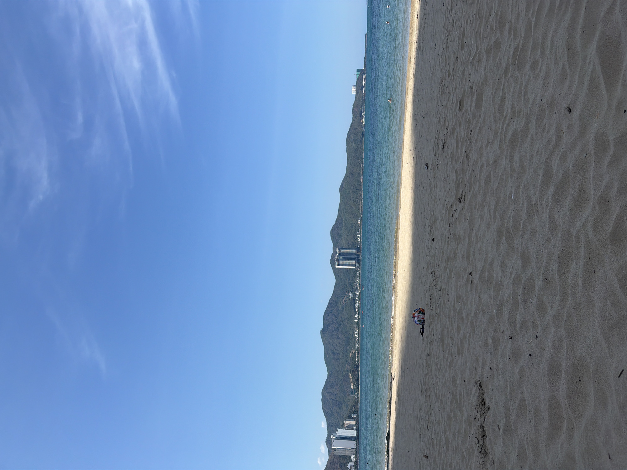 The image shows a beach landscape in Bac Nha Trang Ward, Vietnam. The view is predominantly split between a bright blue sky with wispy clouds and a sandy beach. The water is a turquoise blue, and there are buildings, potentially resorts or residential buildings, visible along the coastline near a mountain range. There appears to be a single person or group relaxing on the beach, evidenced by the presence of a beach umbrella or sunshade. The sand shows patterns of ripples.