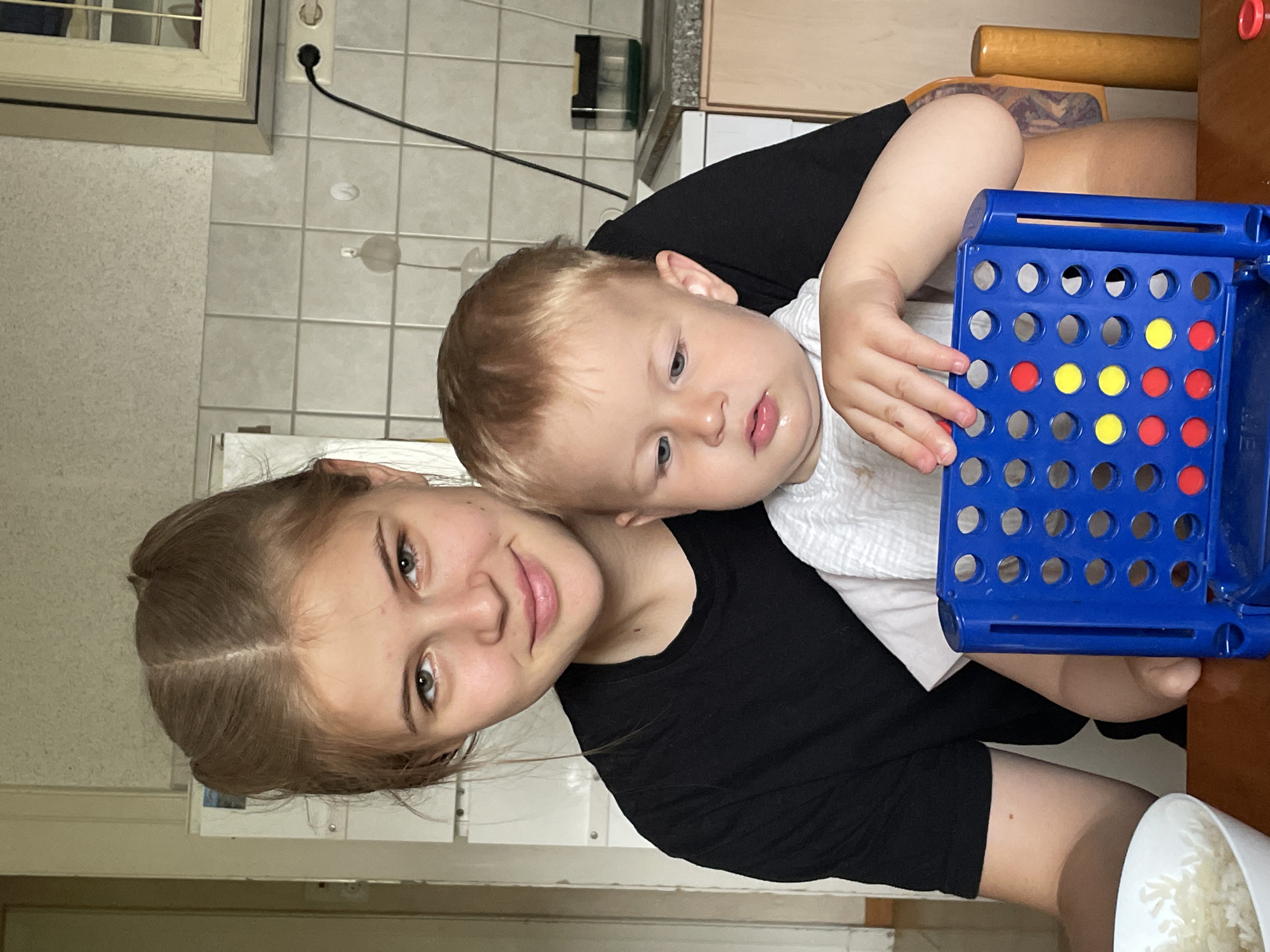 In Kohden, Germany, an image captures a close-up view of a young woman and a baby boy, both sitting at a wooden table. The woman, with light brown hair styled in two pigtails, wears a black t-shirt and is positioned slightly behind and to the left of the baby. The baby, fair-skinned with blonde hair, is wearing a white bib and is seated in front of the woman, partially obscured by her. Both are looking towards the camera.
In the foreground, a blue Connect Four game is visible, with several red and yellow discs already placed. The baby's hands are near the game. To the left of the game, a white bowl containing white rice is partially visible. The background shows a kitchen environment with tiled walls and cabinets.