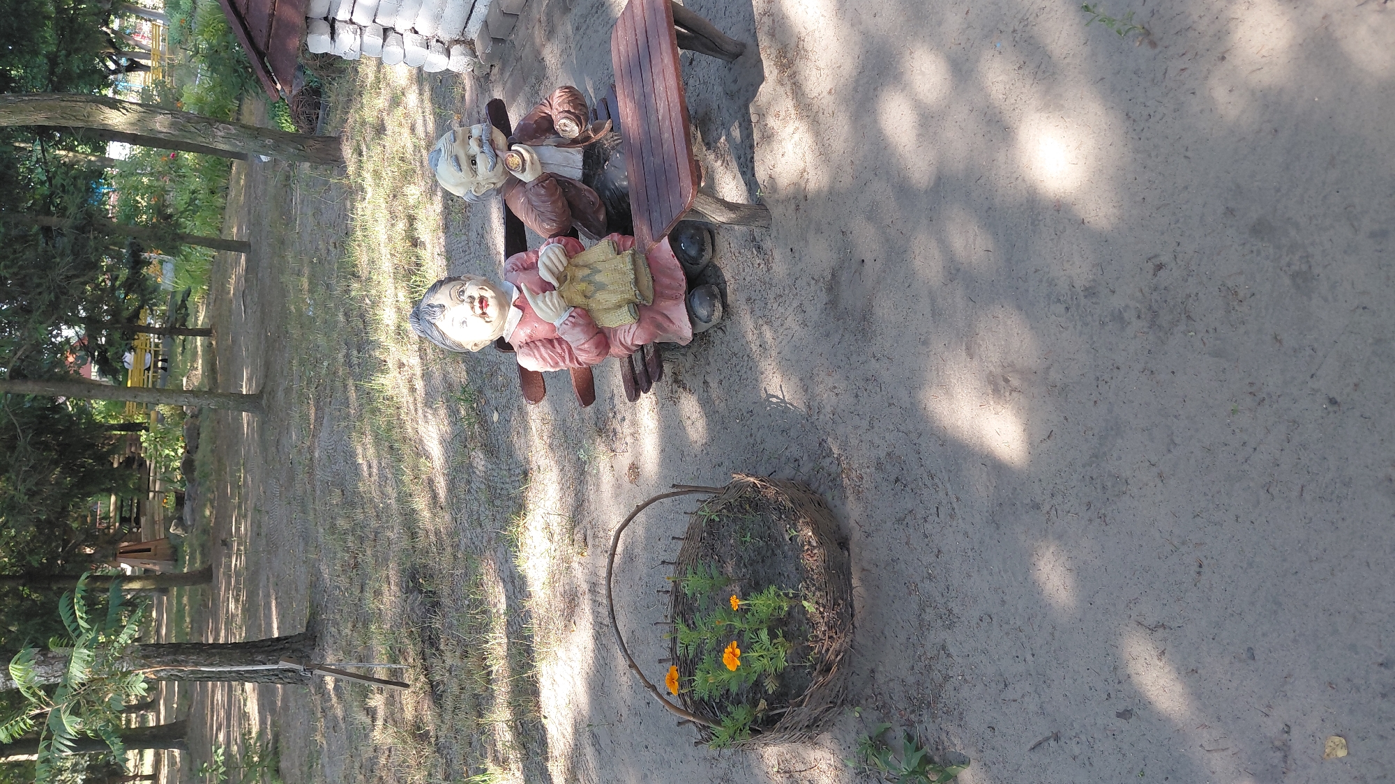 In Borodianka, Ukraine, on a sandy ground with scattered dry grass and tree shadows, a woven basket containing several small orange flowers sits to the left of the frame. To the right, two garden gnomes are seated on a wooden bench. The gnome on the left, a female figure wearing a pink dress and black shoes, is depicted knitting a yellow garment. The gnome on the right, a male figure with a mustache, wears a brown jacket and holds a pipe to his mouth. A small wooden table is positioned in front of the bench. In the background, trees and a wooden structure are visible.