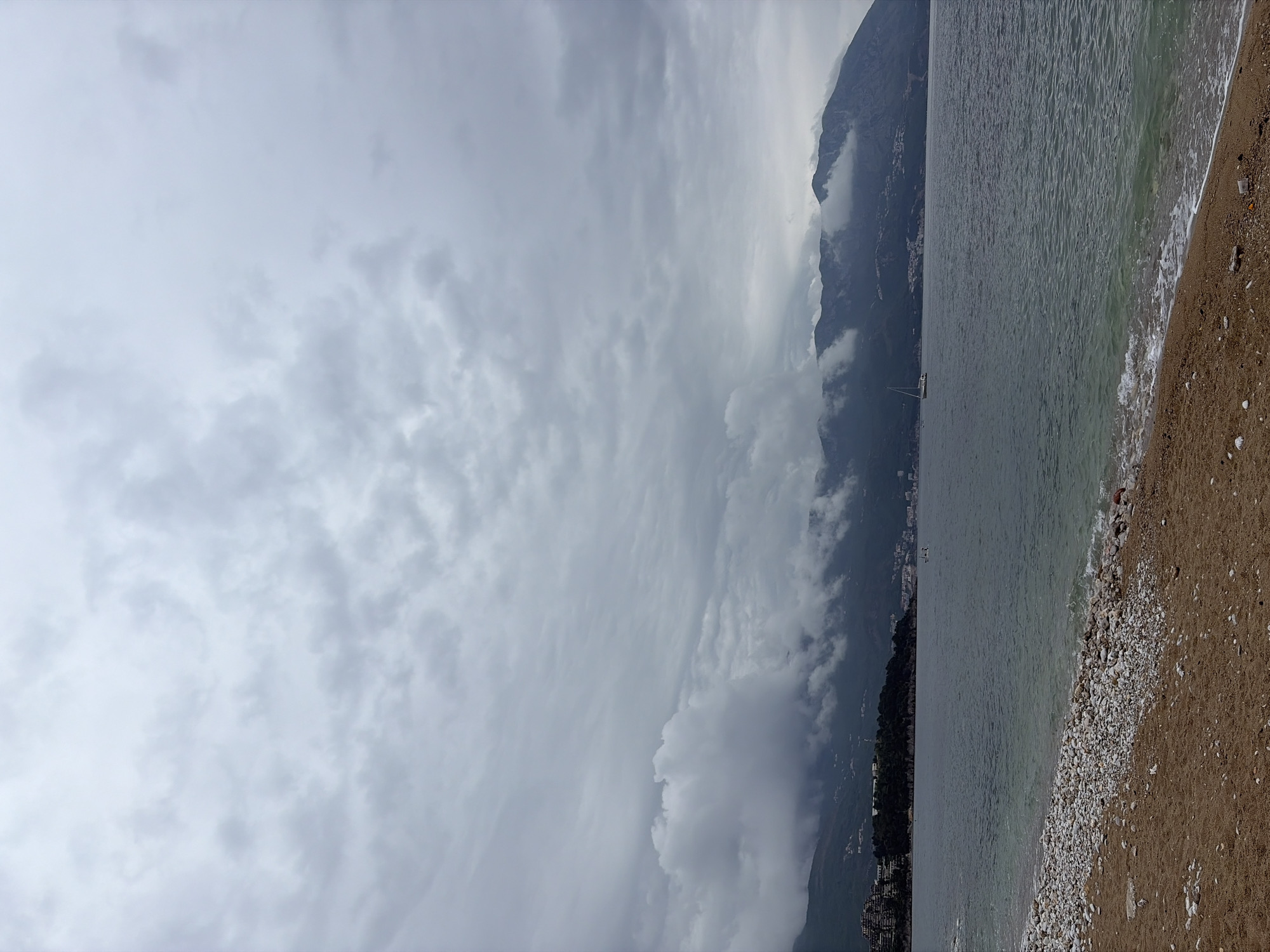 The image shows a cloudy day at a beach in Budva, Montenegro. The view includes the sea, a pebble and sandy beach, mountains partially obscured by clouds, and a gray sky. The water is calm with small waves reaching the shore. No people are visible in the image.