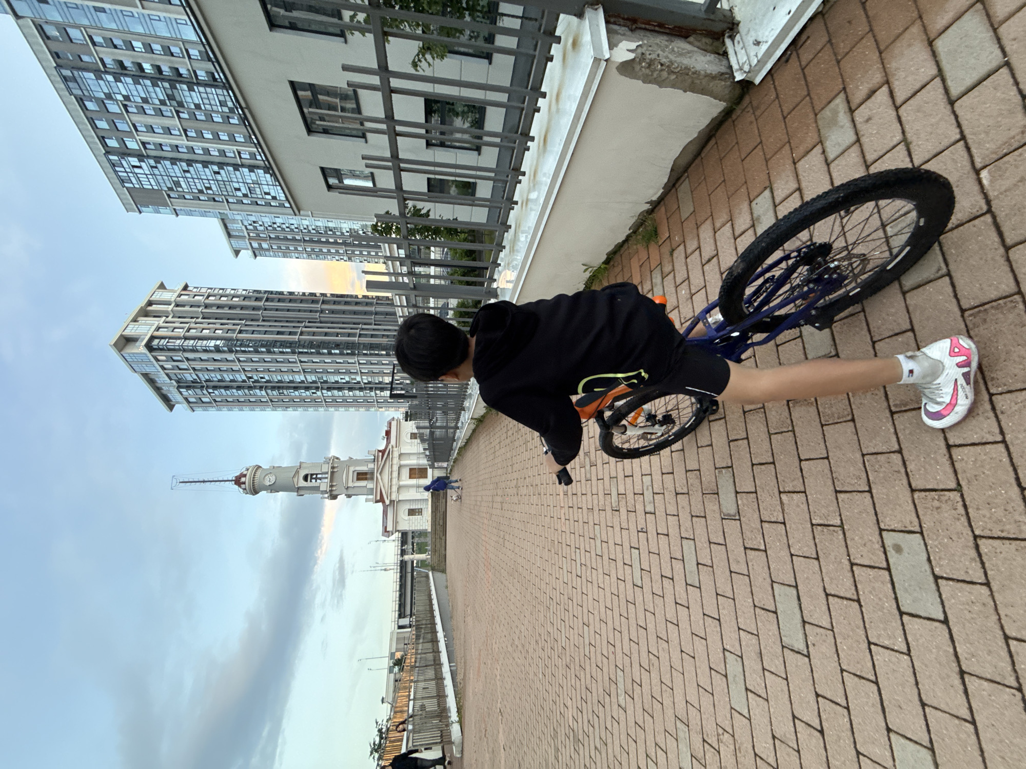 A young boy wearing a black hoodie and black shorts rides a bicycle with an orange frame on a brick-paved walkway. He is viewed from behind, with his right leg extended forward. The bicycle has a blue frame and black tires. The boy wears white sneakers with pink accents. In the background, a white neoclassical building with a clock tower stands under a cloudy sky. To the right of the building, two modern high-rise apartment buildings with many windows are visible. A metal fence runs along the right side of the walkway. A person and a dog are visible in the middle distance, near the white building. Further in the background, a bridge and more high-rise buildings can be seen. The scene appears to be an urban waterfront area.