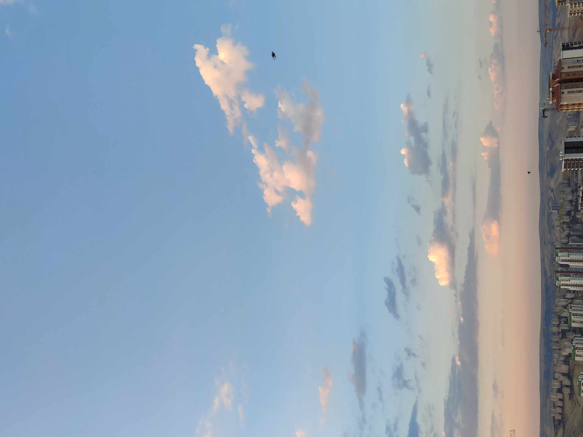 A wide-angle, vertical shot captures a city skyline beneath a vast, pale blue sky with scattered clouds. The clouds are predominantly white, with some tinged with pink and orange, suggesting either sunrise or sunset. A solitary dark bird is visible in flight in the upper right quadrant of the image.
The city itself appears to be a residential area with numerous apartment buildings of varying heights and architectural styles. Some buildings are light-colored, while others feature darker facades. Construction cranes are also present, indicating ongoing development. In the foreground, the closest buildings have a more modern design, with balconies and visible window structures.
The landscape beyond the city is gently rolling hills, some of which appear to be arid or sparsely vegetated. The overall impression is of a modern urban environment nestled within a more natural, undeveloped landscape. The city is identified as Etimesgut, Turkey. There are no discernible people or interactions visible in the image.