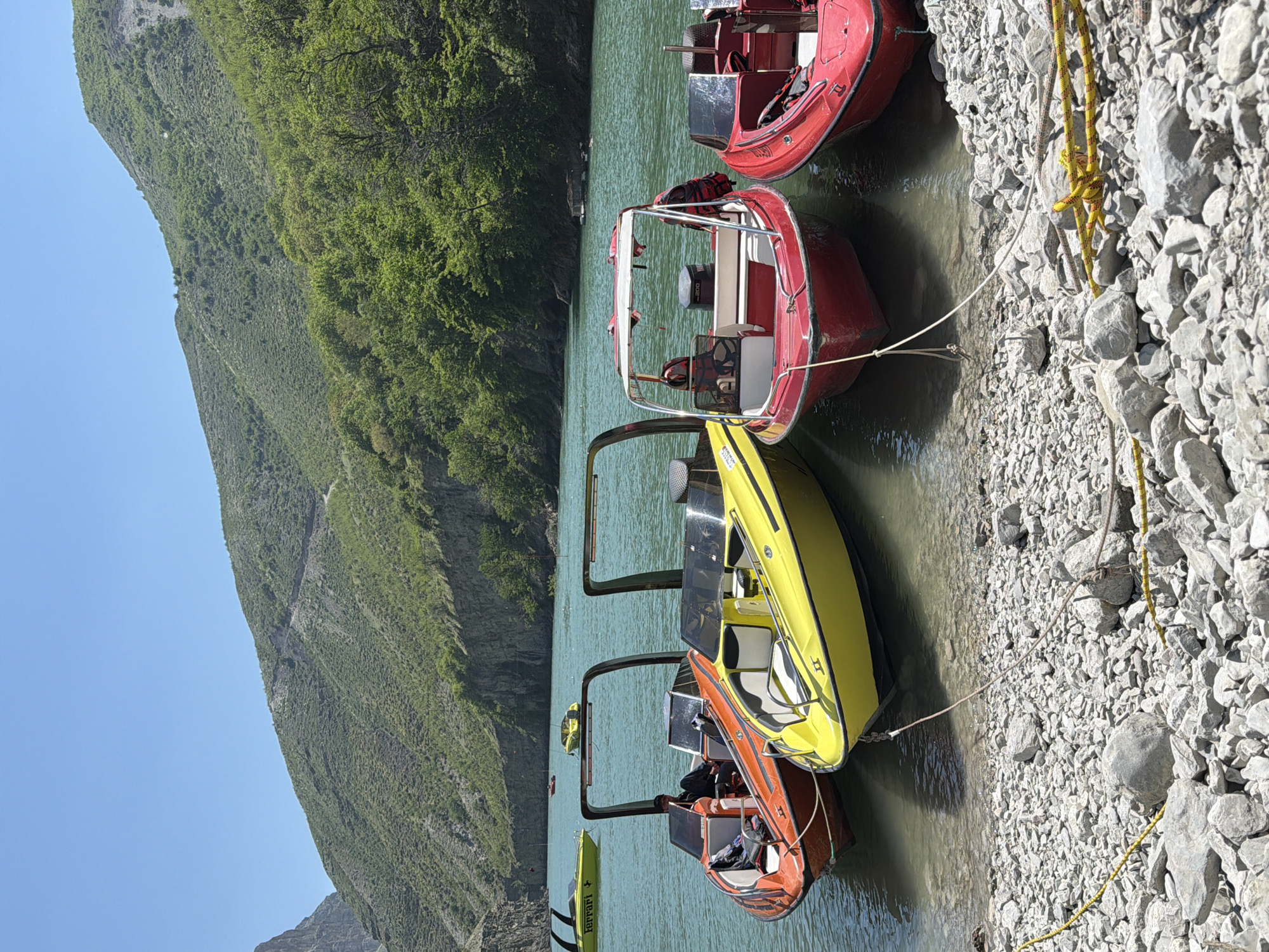 The image shows a view of a lake surrounded by mountains, with several boats docked along the shore. The boats are painted in various colors, including red, yellow, and orange, and are tied to the rocky shoreline with ropes. The water is calm and greenish-blue, reflecting the surrounding mountains and sky. The mountains are covered with lush green vegetation, and the sky is clear and blue. The location is presumed to be in the city of Unknown, Unknown.