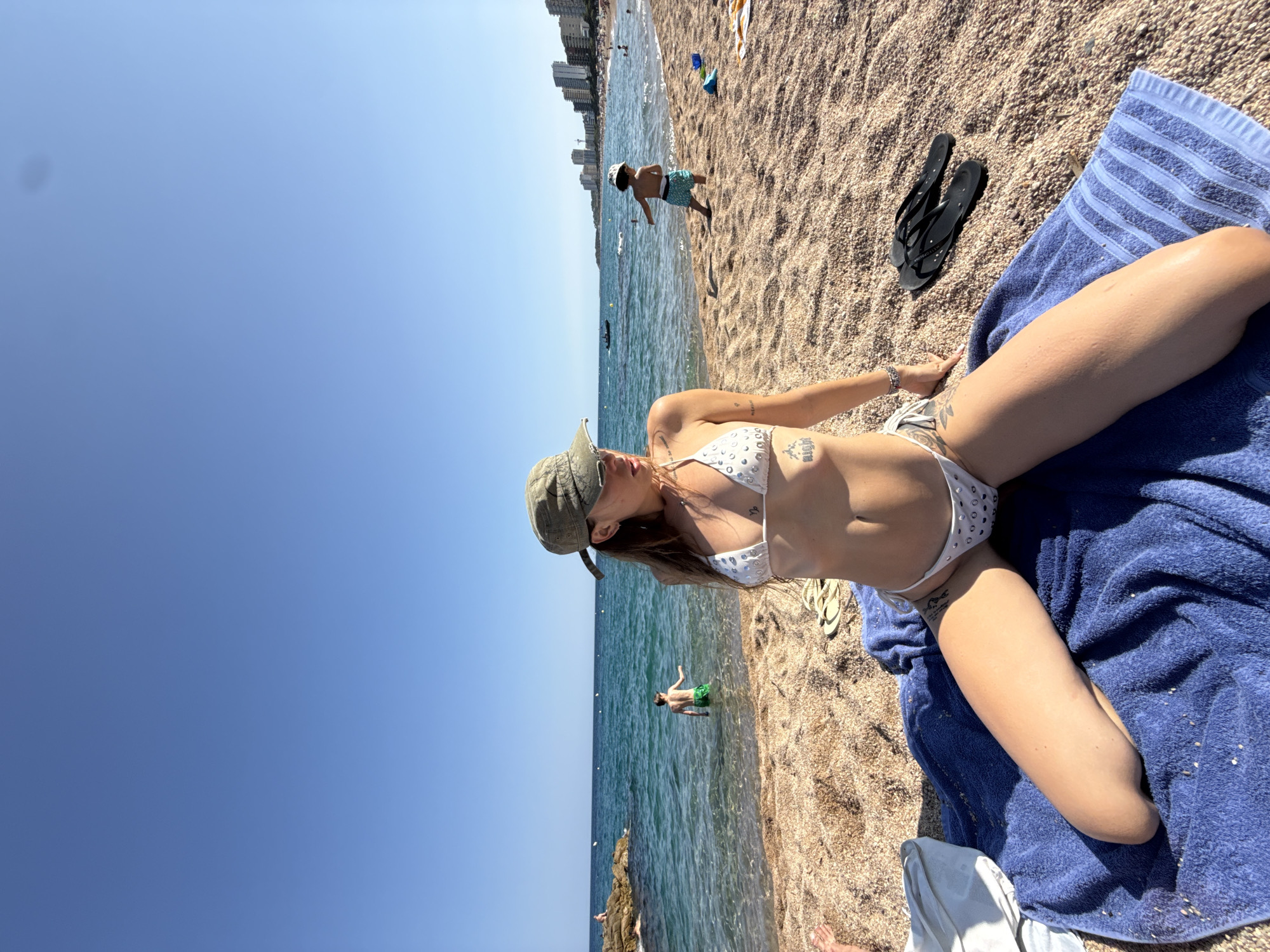 A woman wearing a white bikini and a green cap sits on a blue towel on a beach in Platja d'Aro. She is facing to her right, looking out at the ocean. Her legs are spread apart, and she is resting her arms on her knees. The beach is made of small, light-colored pebbles. In the background, the ocean is visible, with waves gently rolling onto the shore. There are a few people in the water, and further down the beach, there are buildings and a few boats. The sky is clear and blue.