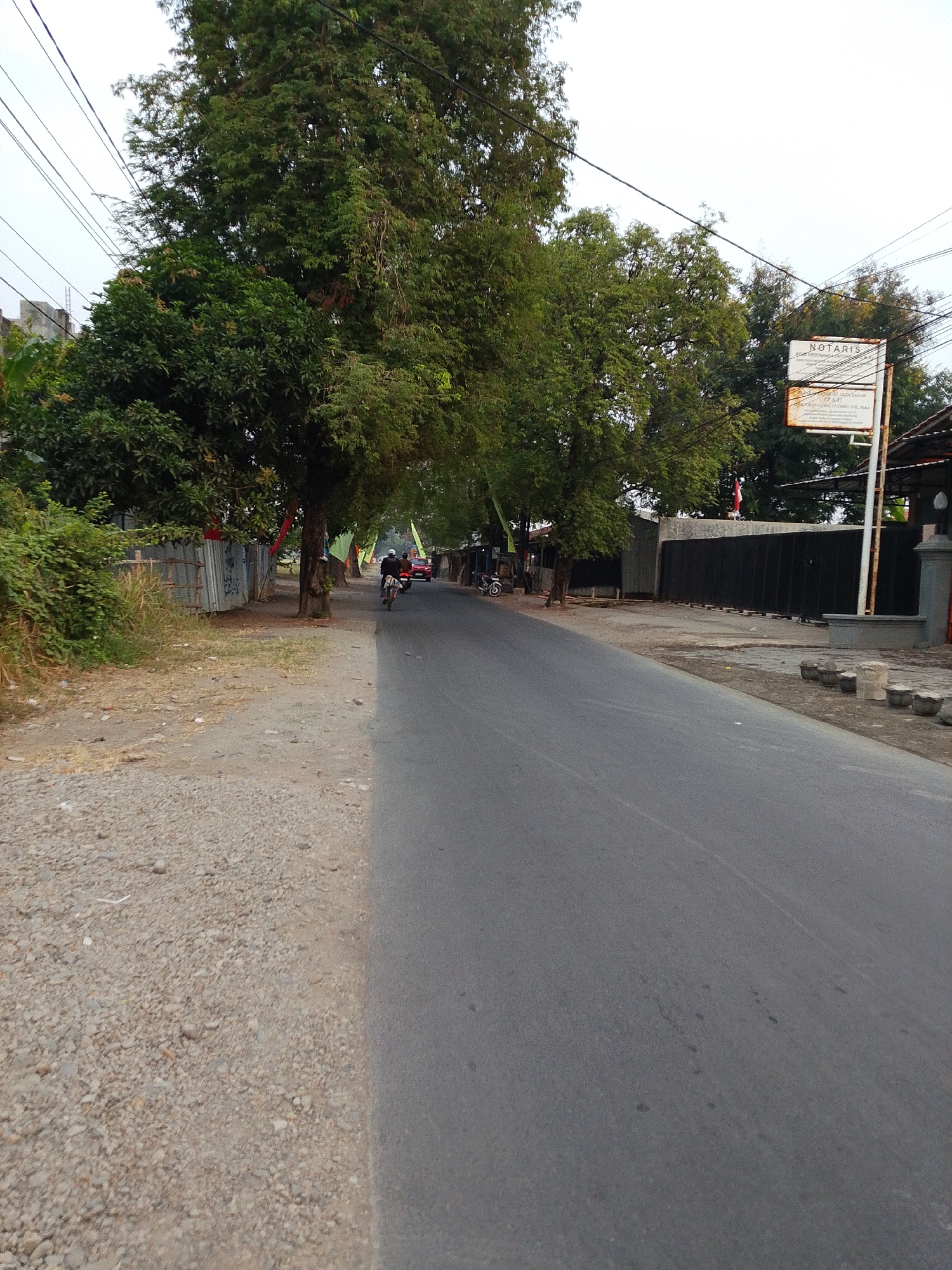 The image depicts a street scene in Tegalwangi, Indonesia. A paved road occupies the right side of the frame, curving slightly to the left. The left side of the road features a gravel shoulder, followed by a dense area of green foliage and trees. In the background, a metal fence and buildings are visible. A signpost stands near the road, displaying text in Indonesian. Several trees with lush green canopies line the street. On the road, a person on a motorcycle is seen riding away from the viewer, and further down the road, a red vehicle is present. The sky is overcast. The overall impression is that of a slightly humid, tropical environment on a cloudy day.
