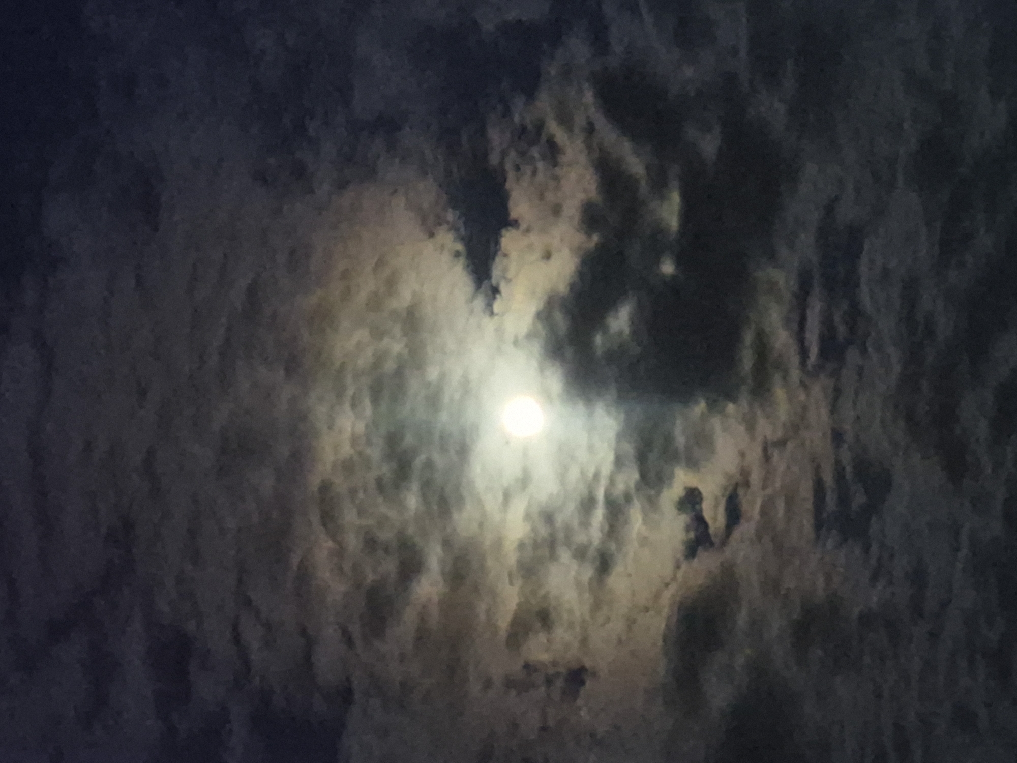 The image presents a striking view of a cloudy night sky illuminated by a bright, diffuse light source, most likely the moon. The central feature is a radiant, circular orb, the moon, positioned slightly above the center, casting a soft, yellowish-white glow. This light penetrates and highlights the surrounding clouds, creating a dramatic contrast.
The clouds are dense and textured, displaying a spectrum of dark grays, deep blues, and lighter white-yellow hues where the moonlight catches them. Some areas of the clouds appear particularly dark and brooding, while others are ethereal and translucent due to the illumination. The overall scene conveys a sense of a heavily clouded night, with the moon attempting to break through the atmospheric cover.
There are no discernible people, objects, or specific location cues beyond the natural phenomenon of the moon and clouds in the night sky. The image has a slightly grainy quality, enhancing its atmospheric and natural aesthetic, evoking feelings of mystery and tranquility associated with moonlit nights. No text is visible.