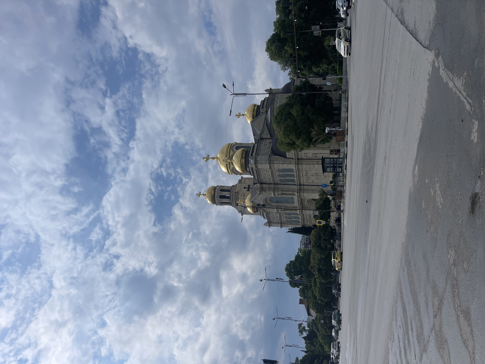 The image displays the Dormition of the Theotokos Cathedral in Varna, Bulgaria, viewed from a broad, wet asphalt street. The cathedral, a large Orthodox Christian edifice, is constructed from light-colored stone with intricate architectural details, featuring multiple golden domes topped with crosses and two taller spires on its left side. Arched windows are visible on its facade.
The street in the foreground shows signs of dampness or recent rain, with visible streaks and reflections on the dark asphalt surface. Several vehicles are present on the road: a white taxi, possibly a Volkswagen Golf, is moving on the right side, and a yellow car is visible further back on the left. Other cars are parked or driving in the mid-ground.
Green trees and dense foliage line the street and surround the base of the cathedral. Streetlights with multiple lamps are positioned along the road, and a traffic signal pole is visible on the far left. Near the cathedral's base, a small bus stop shelter and various road signs, including a prominent yellow diamond-shaped sign, are discernible. The sky above is partly cloudy, featuring patches of blue interspersed with numerous white and grey cumulus clouds. A few indistinct human figures can be observed in the distance near the cathedral and bus stop.