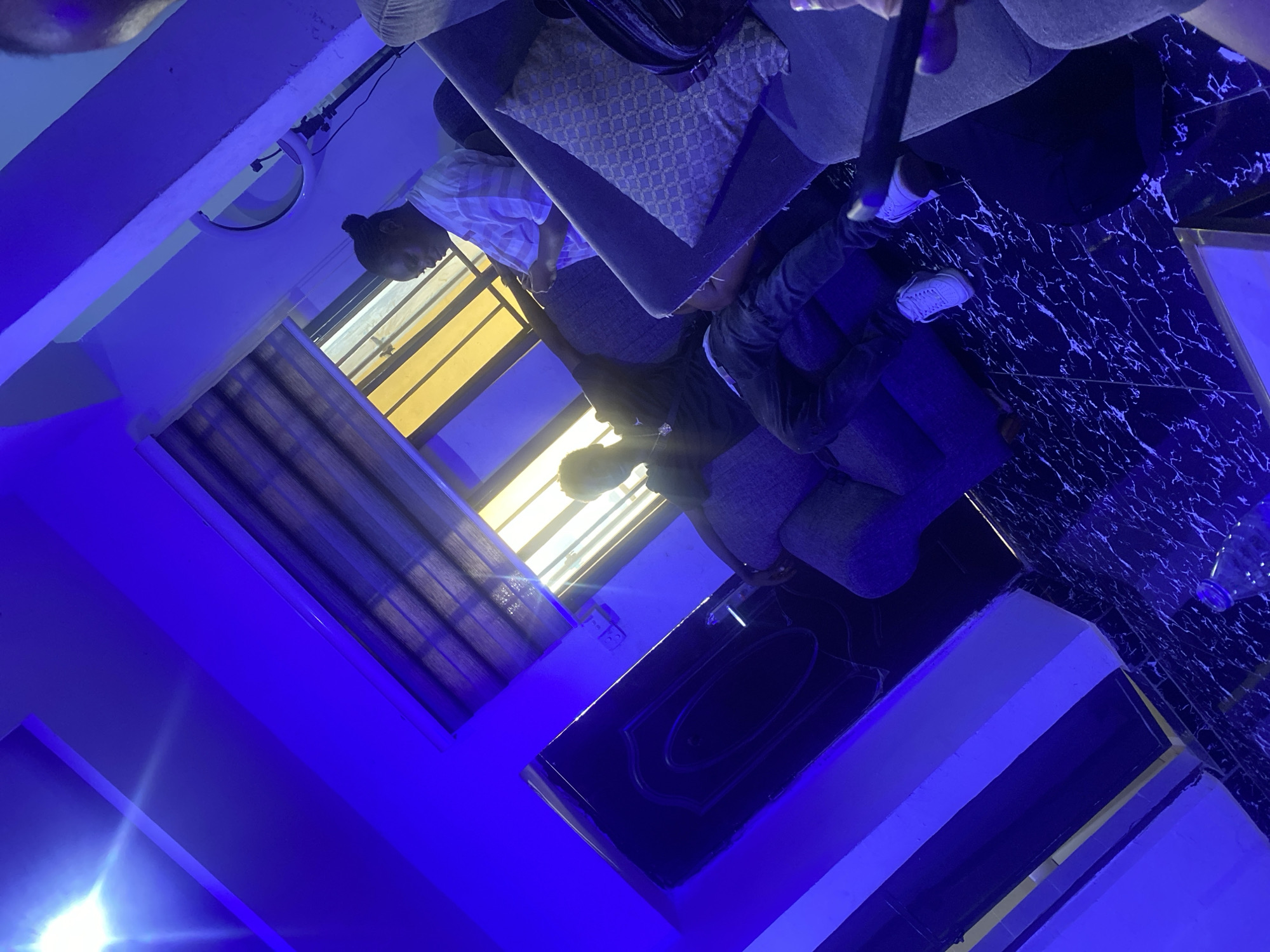 The photo depicts an indoor scene illuminated by blue lighting. The flooring has a black marble pattern. Two individuals are visible; one is partially visible, wearing a light-colored striped shirt. The other individual is wearing a dark jacket. They are positioned near a window covered with blinds and a sofa. A black table is situated near them. The location is in Abuja, Nigeria.