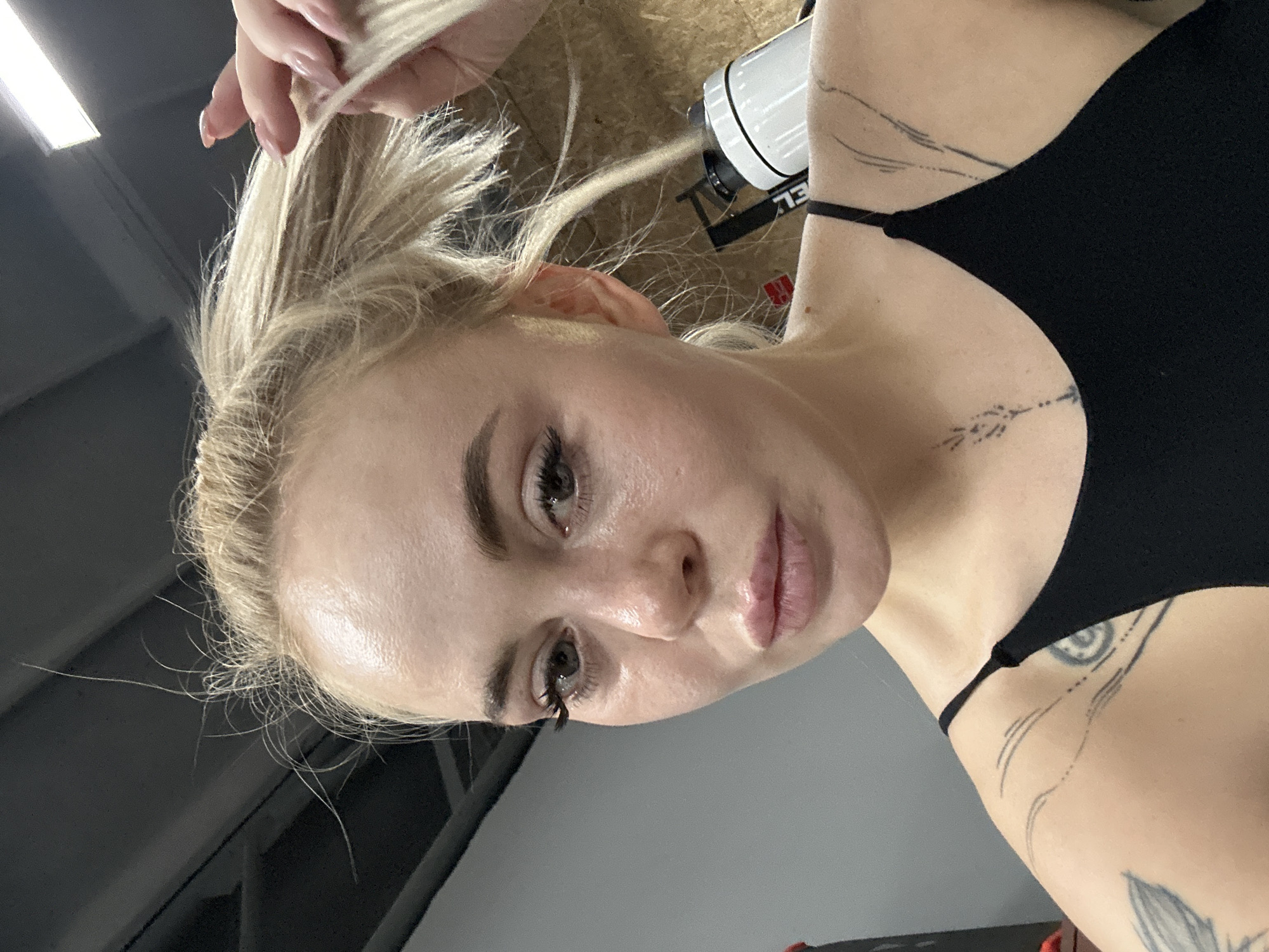 A close-up, angled selfie shows a fair-skinned woman with light blonde hair. Her hair is pulled up, with her hand partially visible, holding strands. She has visible tattoos on her upper chest and shoulder. She wears a black top with thin straps. The background appears to be an indoor space, potentially a gym or studio in Megion, Russia, given the presence of lighting fixtures, ceiling and wall structures.