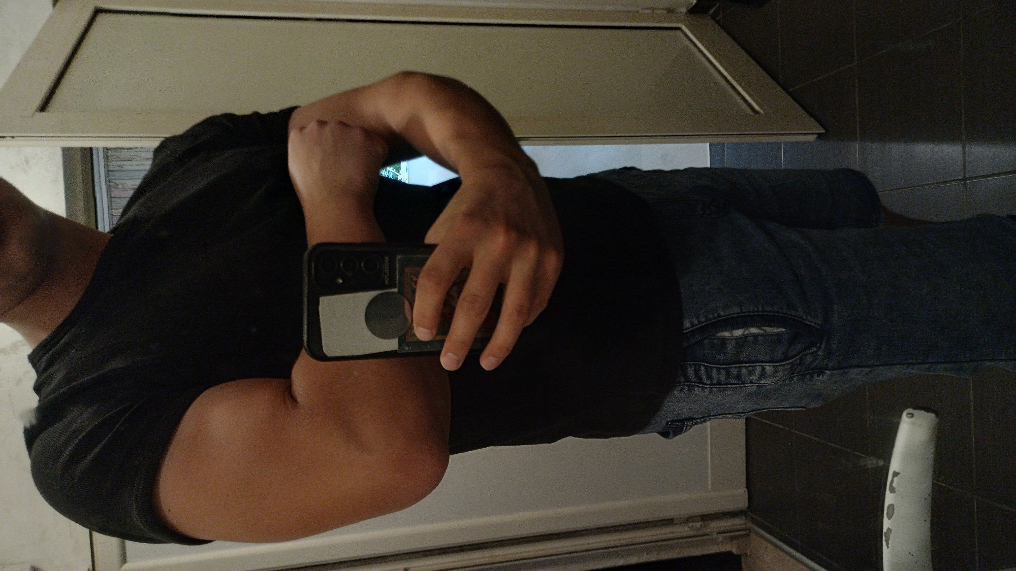 The media file shows a person taking a selfie in a bathroom. The person is wearing a black t-shirt and blue jeans. They are holding a smartphone in their right hand to capture the reflection in a mirror. The background shows bathroom tiles and a white cabinet above the mirror. There is a white pipe or fixture visible in the lower right corner of the image. The lighting suggests it is daytime. The scene is a common bathroom setting.