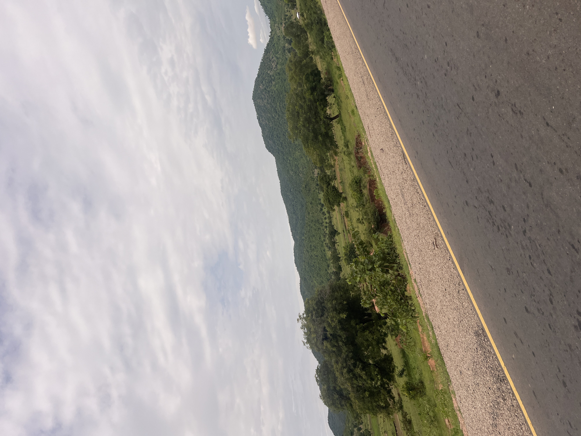 The image captures a scenic view of a highway in Minda, Nigeria. The road, with a distinct yellow line marking its edge, is paved and shows some wear. To the left of the road is a gravel shoulder transitioning into a lush green landscape. Rolling hills covered in dense vegetation dominate the background, suggesting a rural or semi-rural setting. Scattered trees and patches of grass are visible on the slopes of the hills. The sky is partly cloudy, with a mix of white and gray clouds, indicating a potentially overcast or changeable weather condition. The scene appears to be a daytime shot, though the exact time is difficult to ascertain due to the cloud cover. There are no visible people or specific objects that suggest any particular activity or event. The overall impression is one of natural beauty and a tranquil, perhaps remote, location. The angle of the photograph, taken from a slightly elevated perspective and tilted, offers a dynamic view of the road and the surrounding environment.