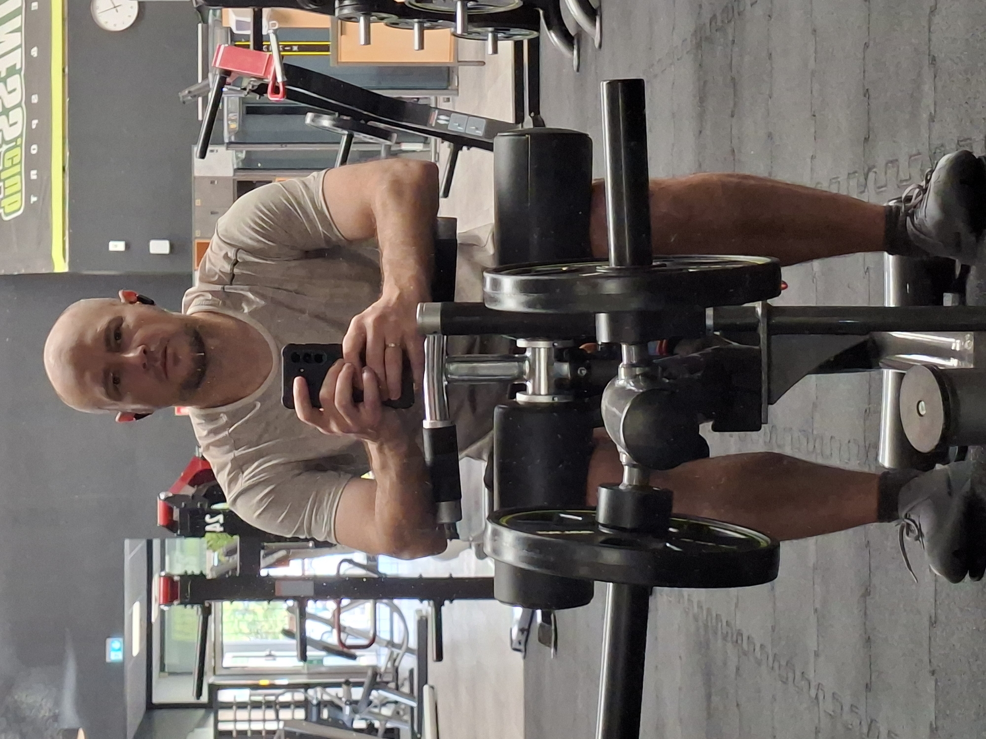 A bald, fair-skinned man with a light beard is taking a mirror selfie in a gym. He is wearing a light-colored t-shirt and dark athletic shorts, with dark grey sneakers and dark socks. He has black wireless earbuds in his ears and is holding a black smartphone in his right hand, positioned to capture the reflection. The gym equipment visible includes weight plates on a machine in the foreground, with other gym equipment and racks in the background. The walls are grey and black, and there is a clock on the wall in the background. The floor is black rubber matting. The overall impression is of a workout session in progress. The image appears to be taken in Baden-Baden, Germany, given the textual elements visible in the background which include "FITNESS CLUB SPORT".