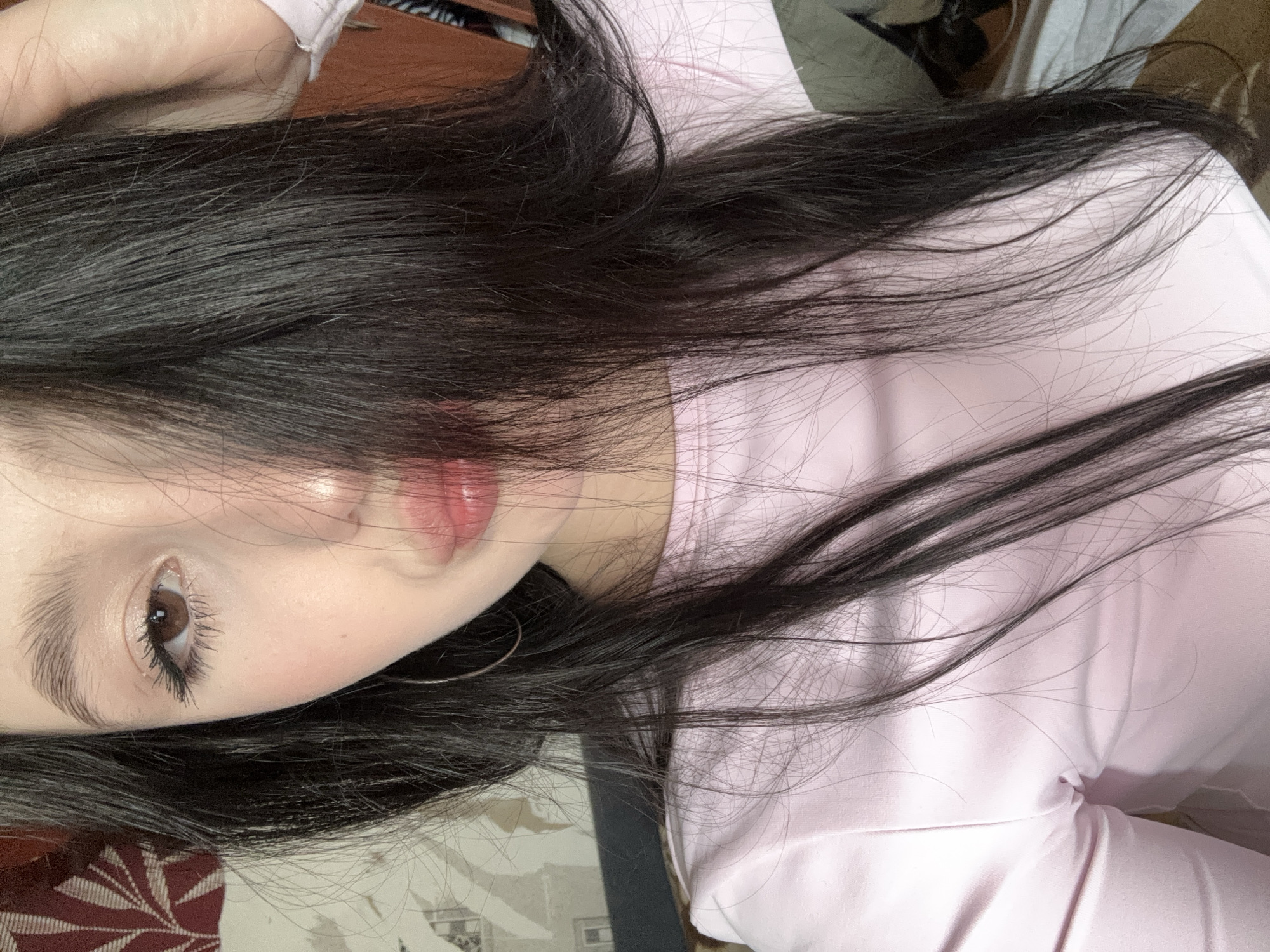 The image depicts a female subject in a close-up, vertical portrait taken indoors, an unspecified location within the city of Фоки, Russia, as per external context. The subject has fair skin, a visible dark brown right eye with mascara-enhanced eyelashes, and a prominent dark eyebrow. Her lips are pursed into a pout and exhibit a glossy light pink coloration. Long, dark hair covers the left side of her face and central facial features. A thin silver hoop earring is visible in her right ear. She is wearing a light pink, long-sleeved garment. The background consists of a reddish-brown patterned textile or wallpaper in the upper left, transitioning to a lighter, subtly textured wall surface below, with indistinct darker elements to the right.