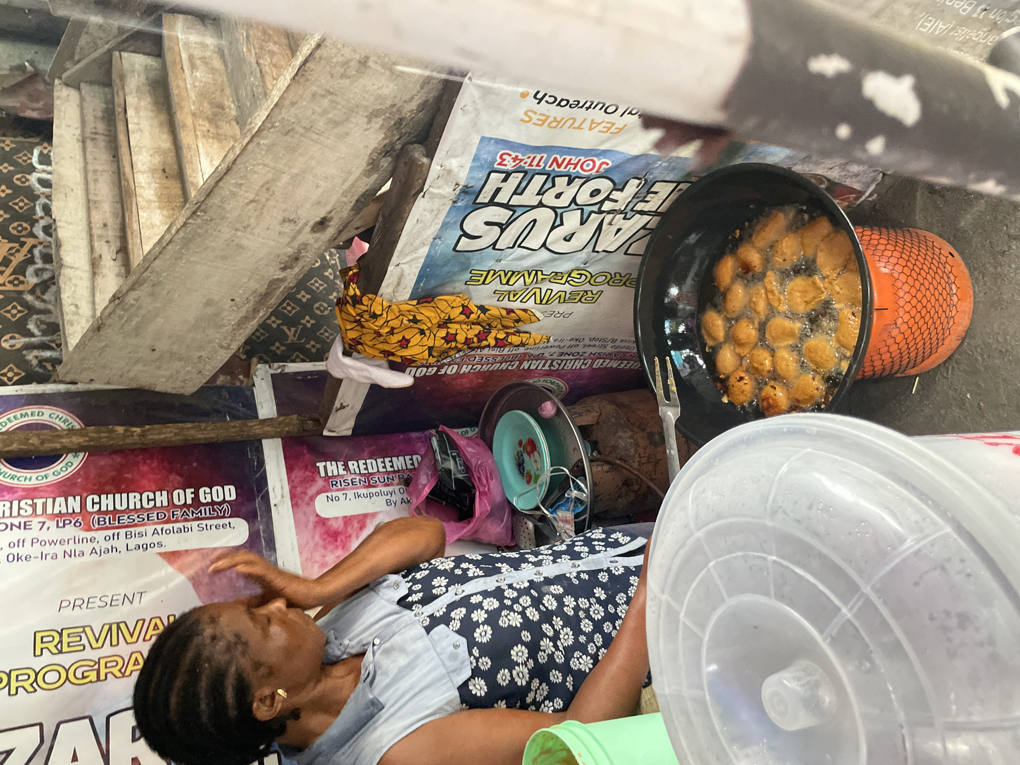 A woman is visible on a densely packed street in Ado, Nigeria, surrounded by signs advertising The Redeemed Christian Church of God. She is lying down, positioned amongst a stack of plates, a pink plastic bag, and a large plastic container. Next to her, a pot of what appears to be frying food is placed atop a bright orange gas cylinder. Adjacent to this cooking setup are rough, wooden stairs, and promotional posters for revival programs at the church.