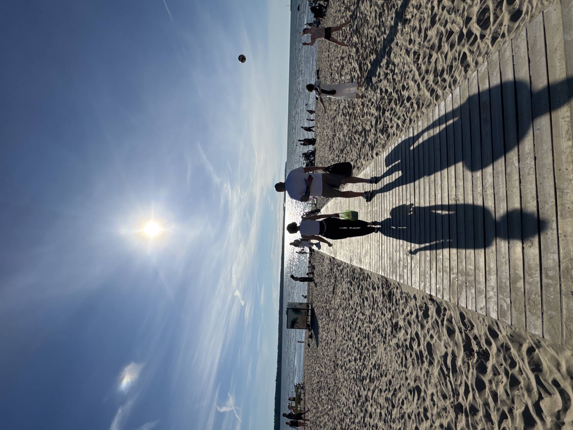 The image depicts a sunny day at a beach in Tallinn, Estonia. A wooden boardwalk extends into the sand, leading towards the sea. Two figures, a man and a woman, are walking away from the camera on the boardwalk, their long shadows cast on the planks. The woman is wearing black pants and a white top, carrying a green bag. The man is wearing shorts and a white t-shirt, carrying a black bag.
In the background, the sea is visible, with people scattered across the sandy beach. Some individuals are playing volleyball, with a ball in mid-air. Other people are seen walking, sitting, or standing near the water. In the distance, a long line of trees borders the coastline, and the sky is a clear blue with wispy clouds and the bright sun shining. The overall scene conveys a relaxed atmosphere of a summer day at the beach.