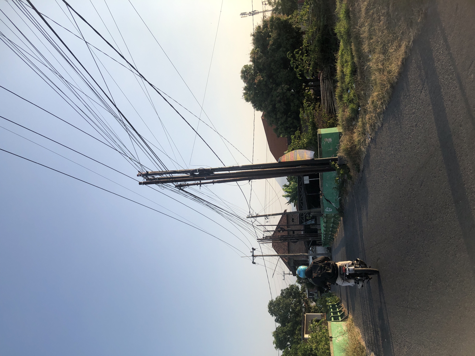 A person rides a motorcycle down a paved road in Kalidawir, Indonesia. The rider is wearing a blue helmet and a dark jacket. The road is bordered by green metal railings and dry grass. In the background, there are several buildings with red tile roofs, lush green trees, and a clear blue sky. Numerous power lines crisscross the sky, supported by wooden poles. The scene is bathed in the warm light of what appears to be either early morning or late afternoon.
