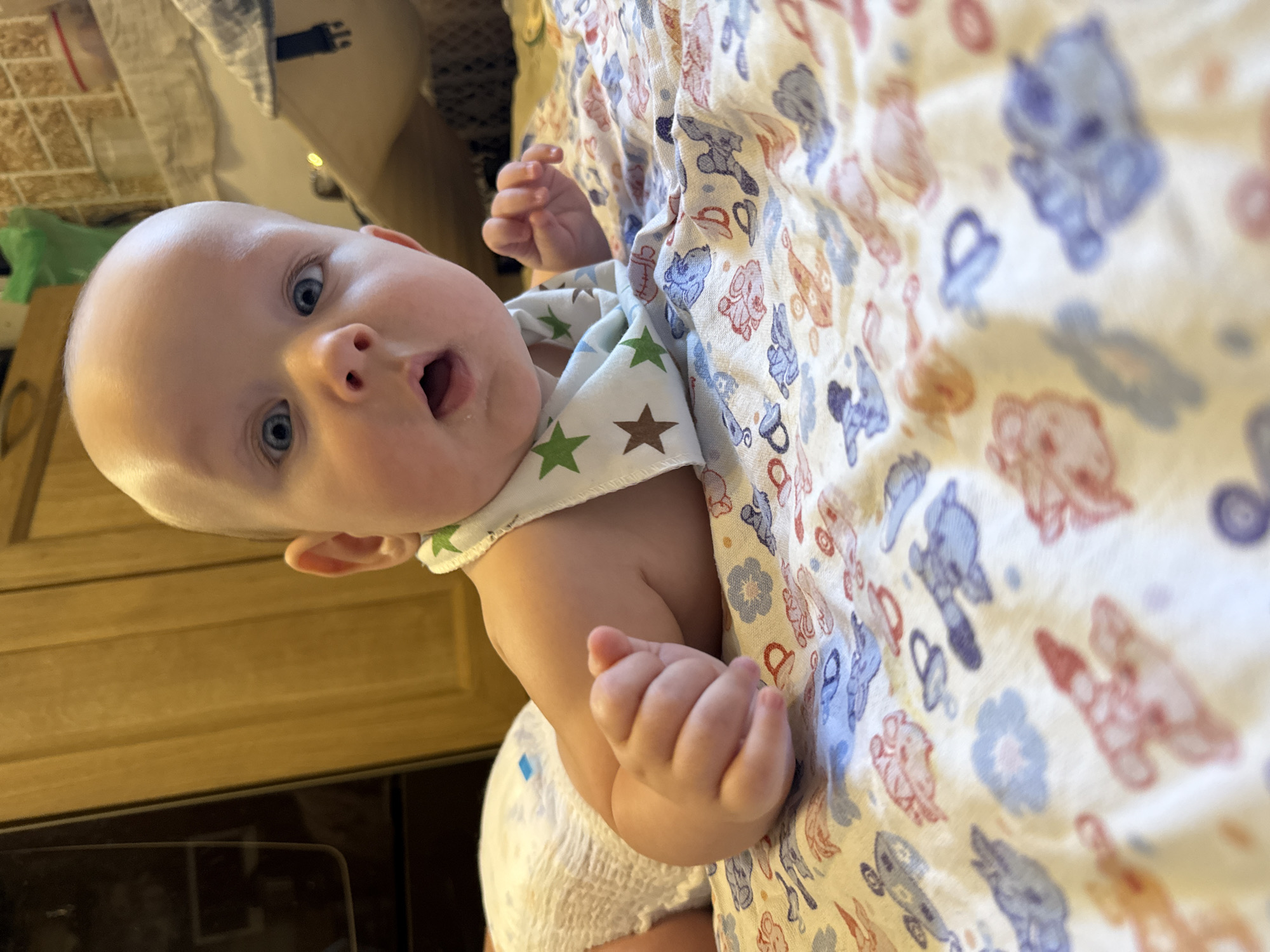 An infant, with a bald head and blue eyes, lies on its stomach on a patterned blanket. The infant wears a bib with green and brown stars and a diaper. The background consists of wooden cabinets and kitchen counter with some items on it. The image is taken from a low angle, with the infant's face as the central focus. The location is an indoor setting, likely a home kitchen, in Moscow, Russia.