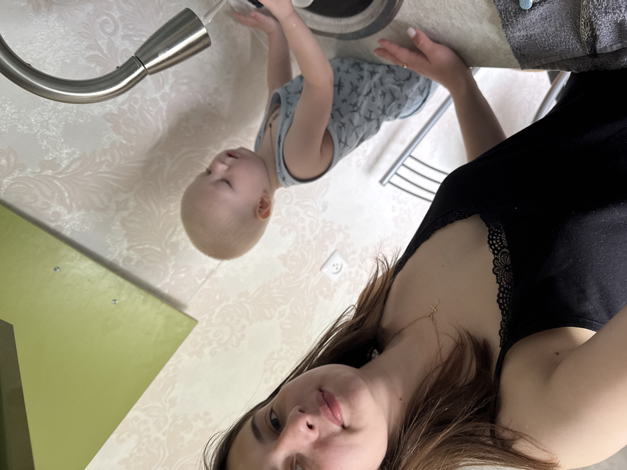 The image depicts a selfie taken indoors. In the foreground, a woman with brown hair is visible, wearing a black top with lace detail around the neckline and a gold necklace with a cross pendant. Behind her, a young child in a gray tank top with airplane patterns is reaching for water flowing from a stainless steel faucet into a sink. The sink is surrounded by a patterned white wallpaper. The area also includes a green-colored element, potentially part of a cabinet or wall. The presence of the faucet and sink suggests the location is a kitchen or bathroom. The image doesn't provide enough detail to determine its specific location within Chaikovsky, Russia.