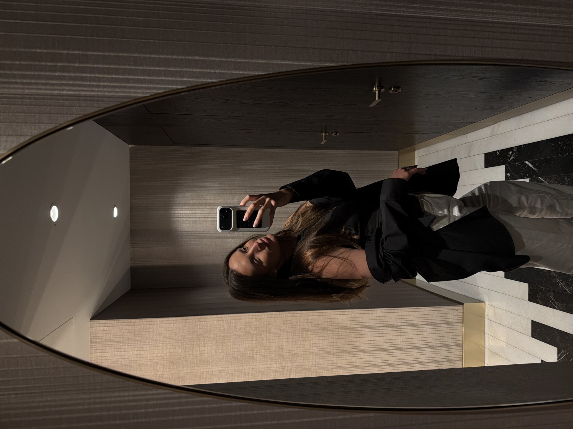 A woman takes a selfie in an oval mirror. The mirror reflects a corridor with wood-paneled walls, round recessed lighting, and a dark wood ceiling with gold-toned fixtures. The floor is white and black marble with gold-toned trim. The image was taken in Budva, Montenegro.