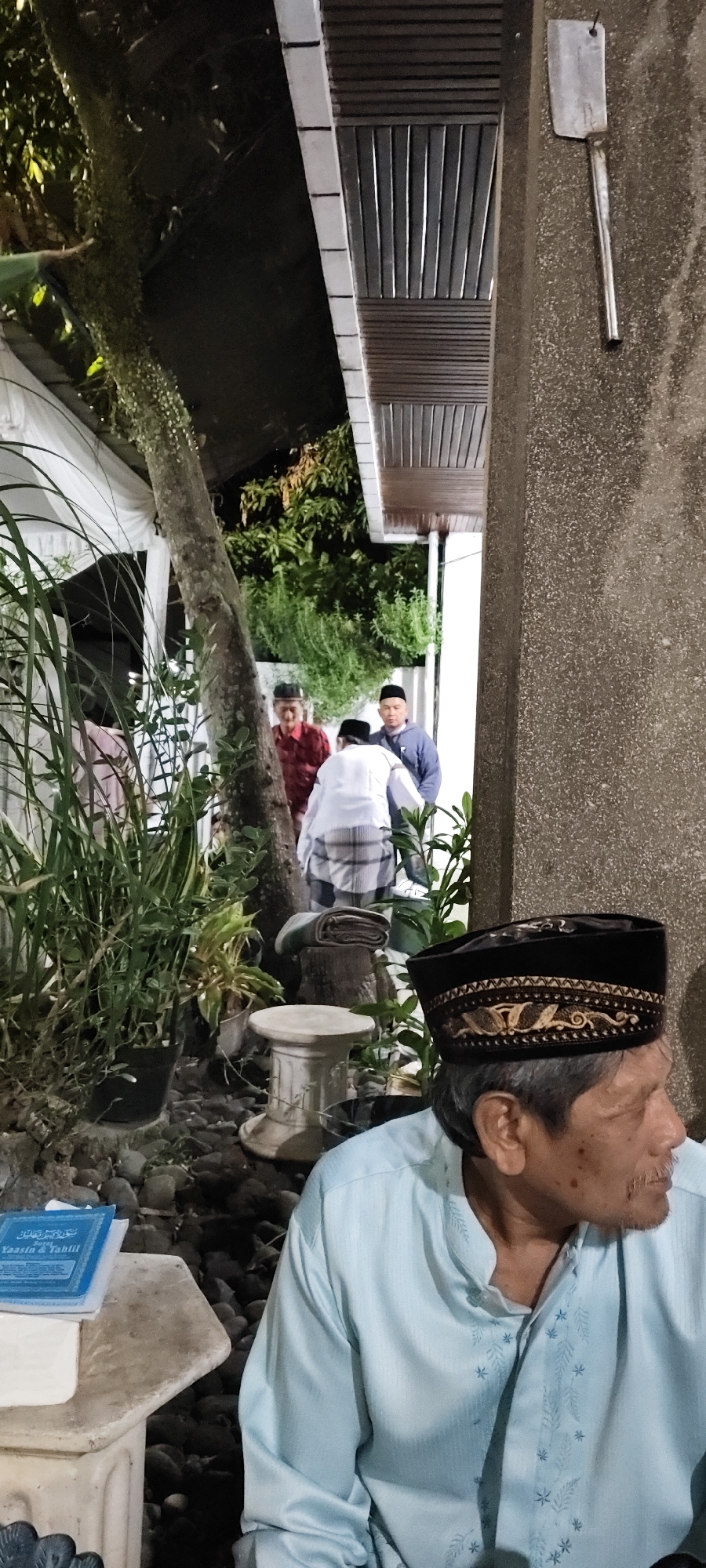 The image depicts a gathering of several men outdoors at night, likely in a residential area in Malang, Indonesia. In the foreground, an elderly man with a dark, embroidered peci (a cap) sits in profile, looking to the right. He is wearing a light blue shirt with subtle embroidered detailing. Behind him, to the left, are lush green plants in pots, and a tree trunk with moss and vines. In the midground, three men are visible. One man in a red shirt stands partially obscured by a tree. Another man in a white garment, possibly a sarong and a shirt, is bending slightly. A third man in a blue jacket and a peci stands behind him. The background shows a building with wooden louvers under a roof, and more foliage. To the right of the building, a concrete wall is visible with a metal cleaver hanging on it. The ground is covered with stones and gravel. A small, ornate stand or pedestal is situated between the plants and the bending man. A blue book, likely religious text given the context of the peci caps, rests on a white surface in the lower left corner. The lighting suggests artificial illumination from the property, casting shadows and highlighting certain areas.